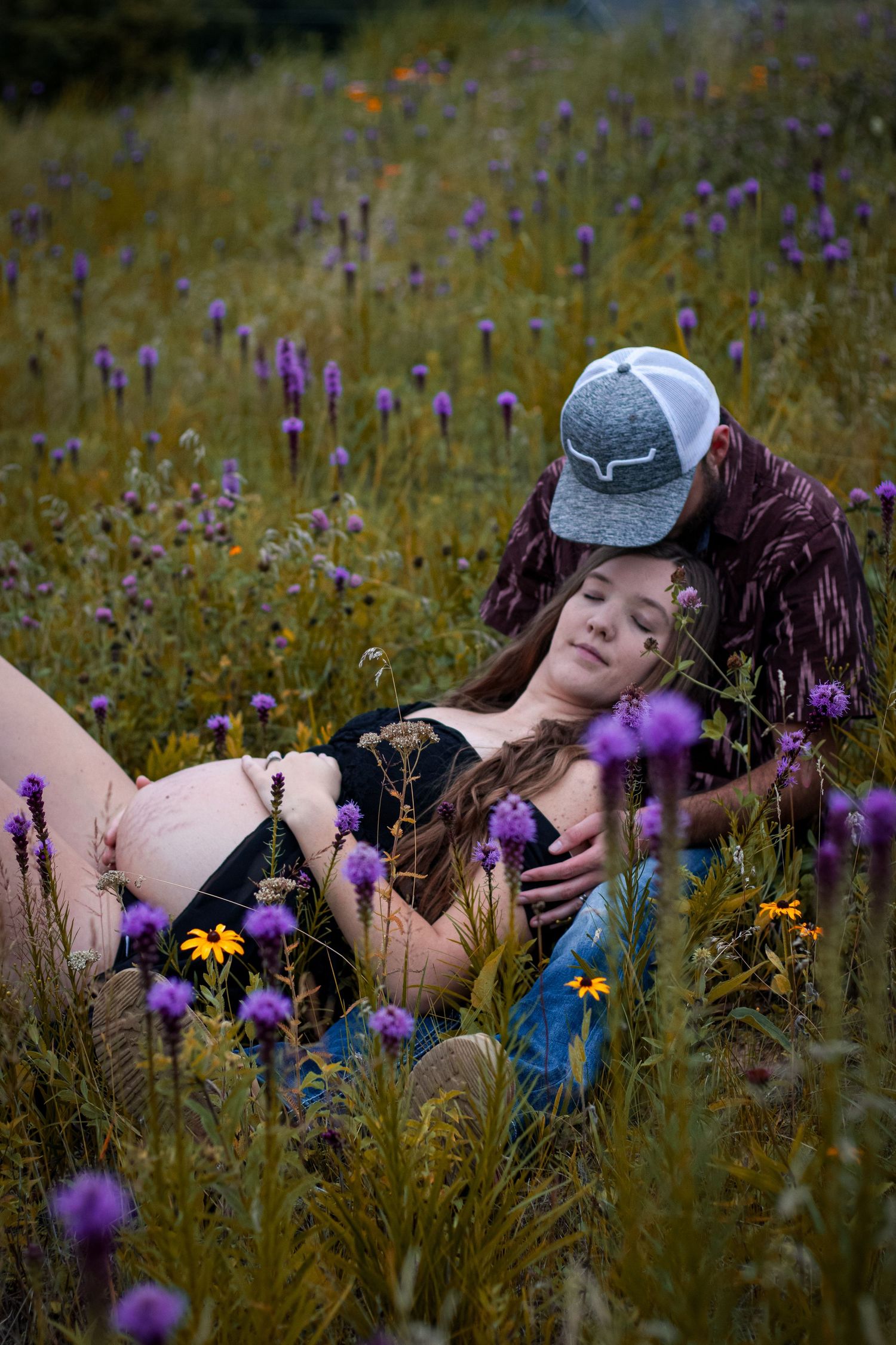 Expecting couple relaxes together in a wildflower meadow filled with purple and yellow blooms.