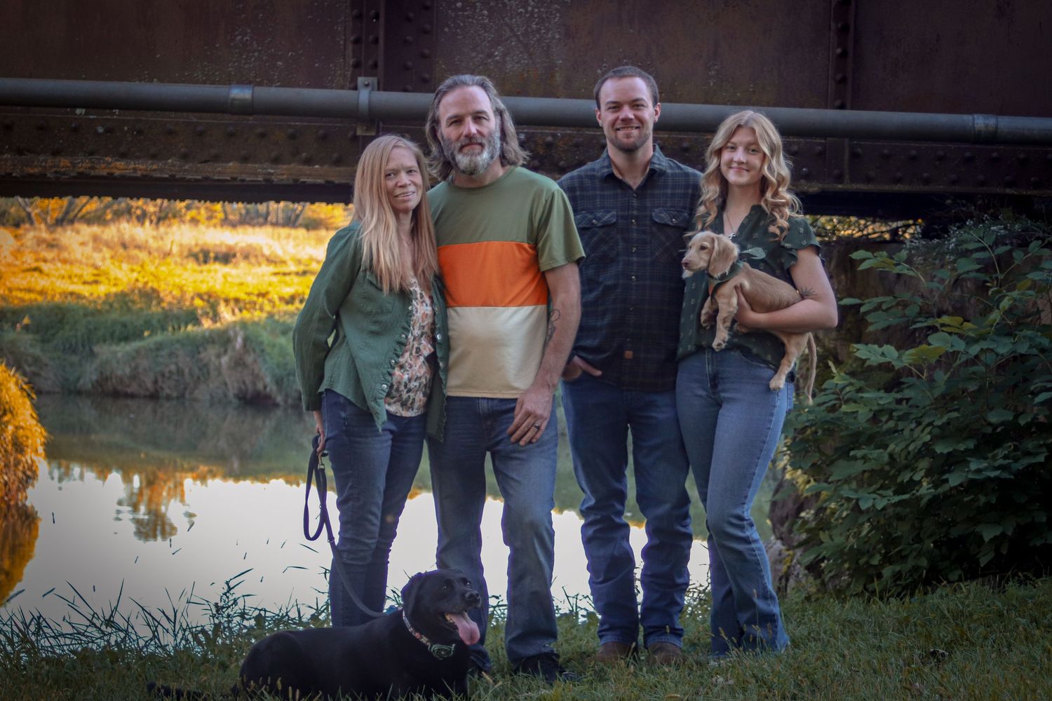 Two couples posing near a river with a bridge backdrop, accompanied by two dogs.