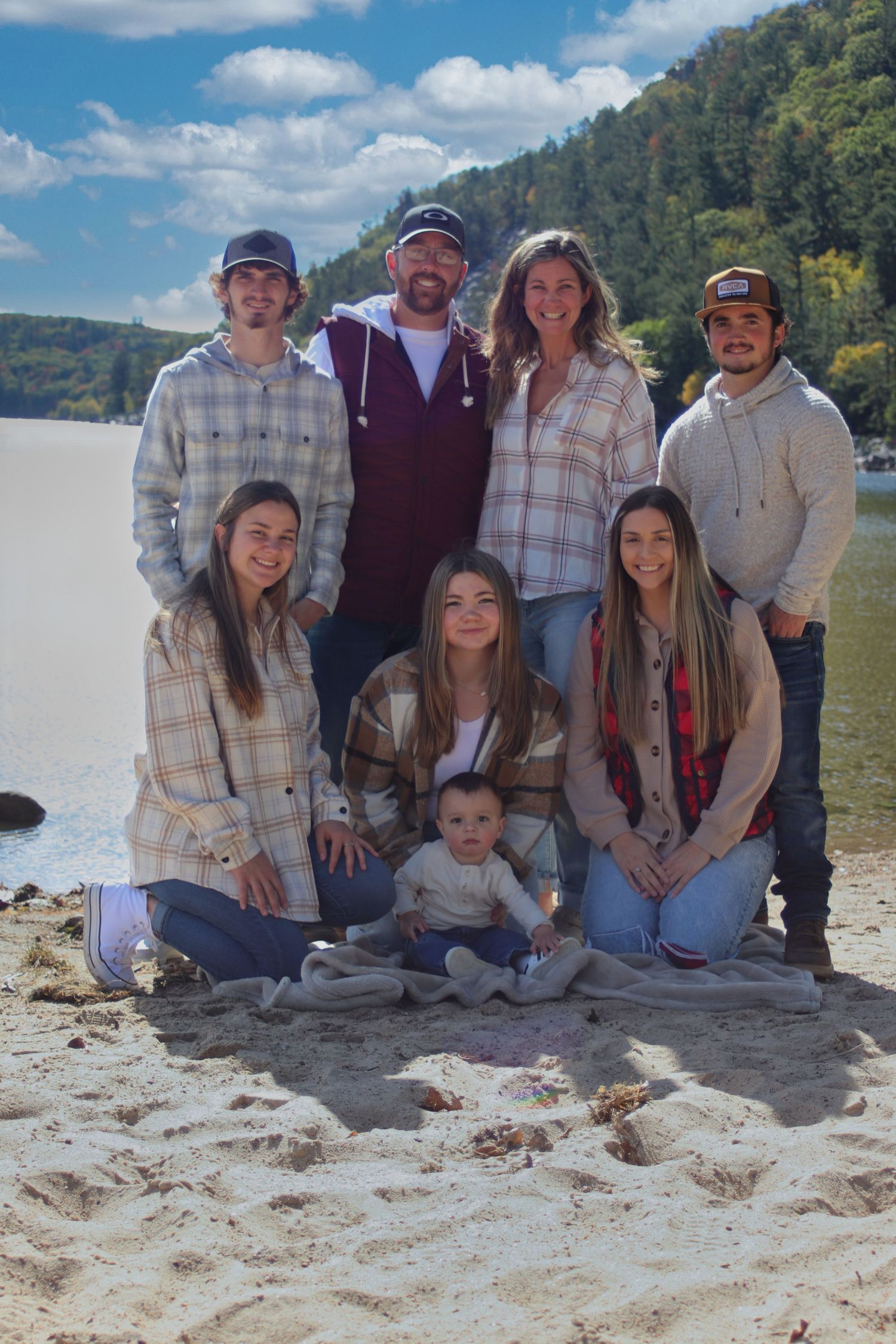 Family of eight posing on a sandy beach with a scenic lake and hills behind them.