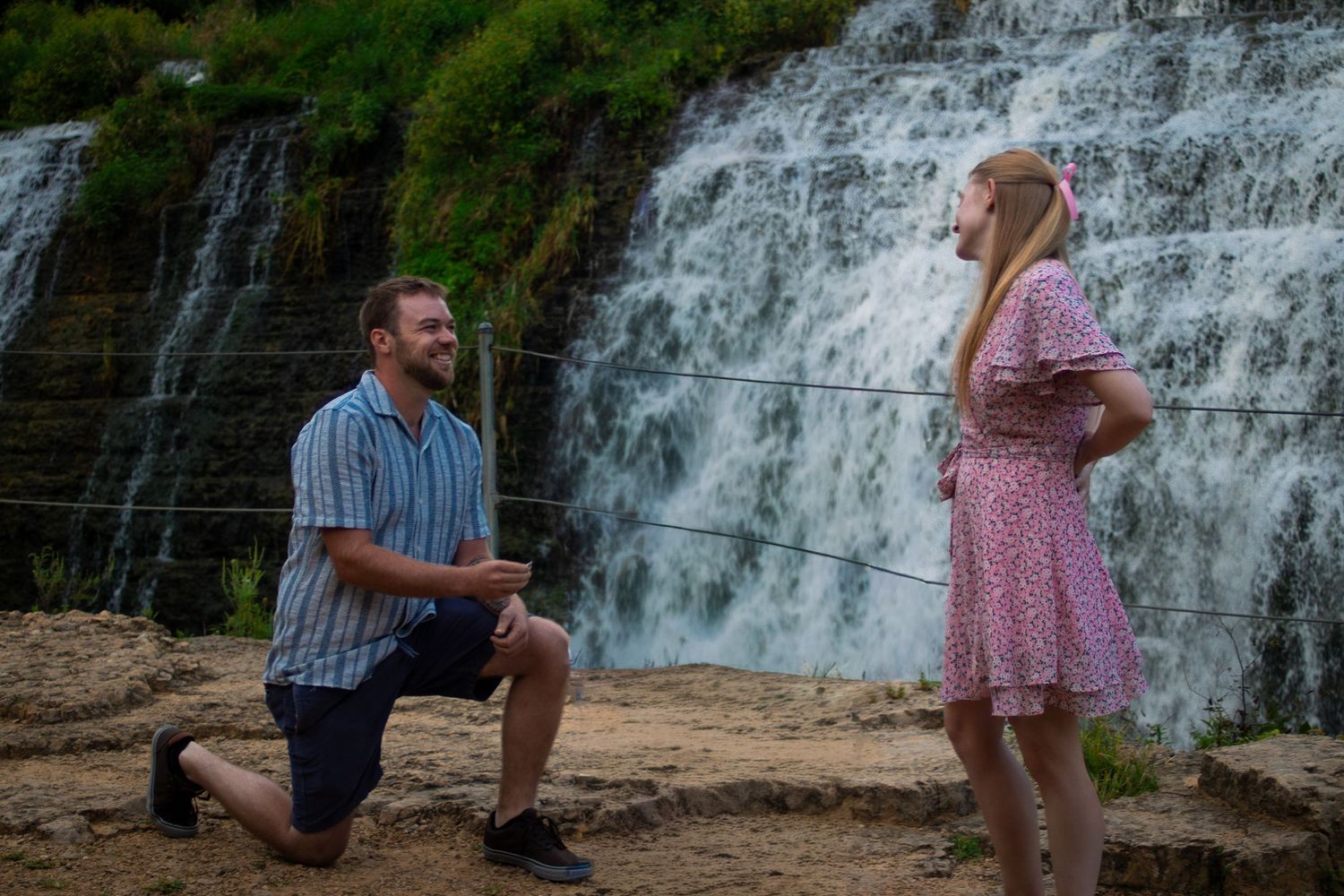 Man proposing on one knee to girlfriend in pink dress in front of scenic waterfall.