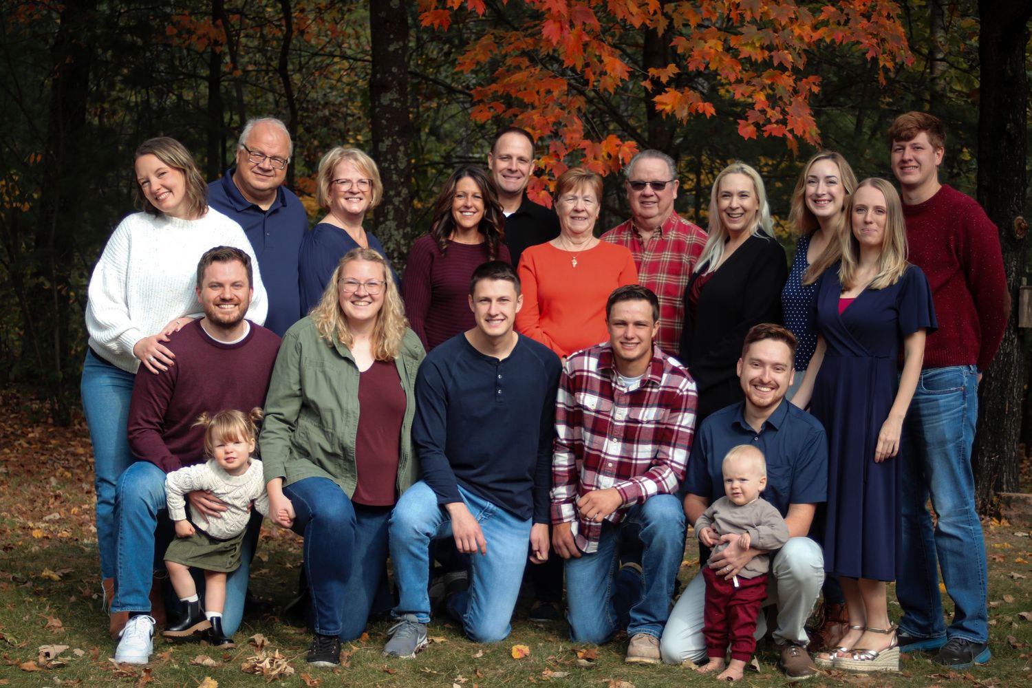 Large multigenerational family poses together outdoors surrounded by vibrant autumn foliage for group portrait.