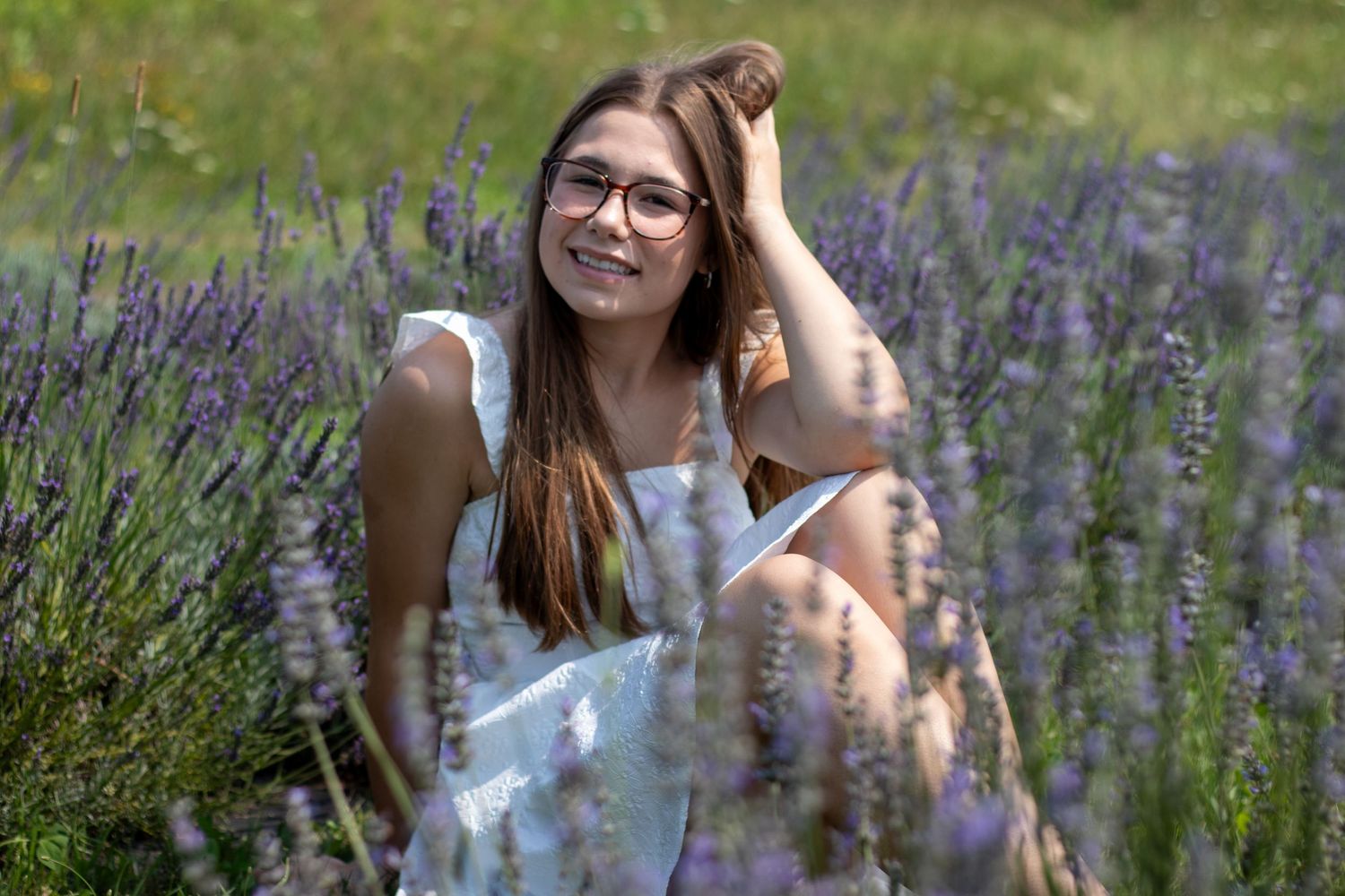 Smiling teenage girl in white dress and glasses sits among purple lavender flowers for senior portrait.