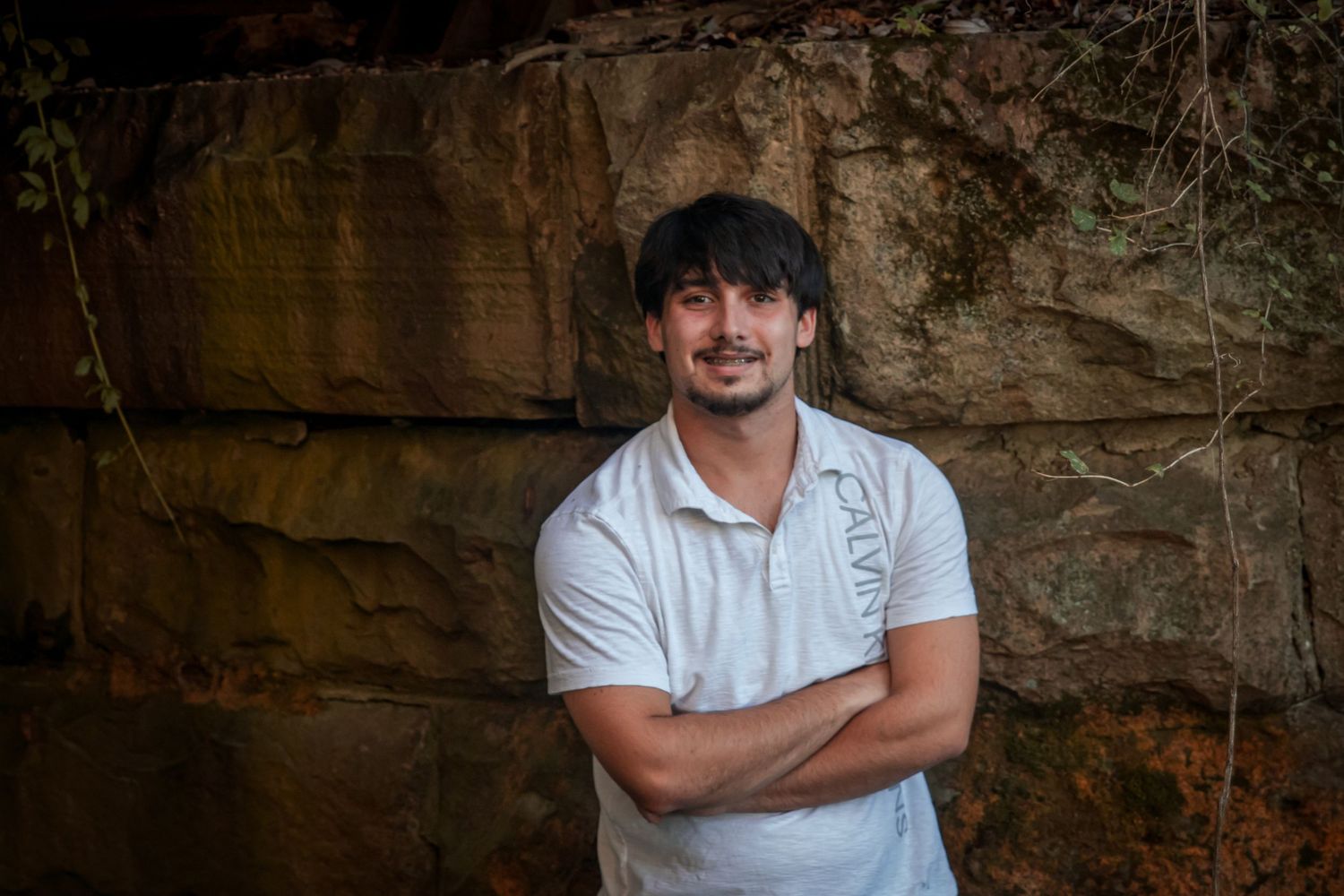Young man with crossed arms smiling in front of a rustic sandstone rock wall.