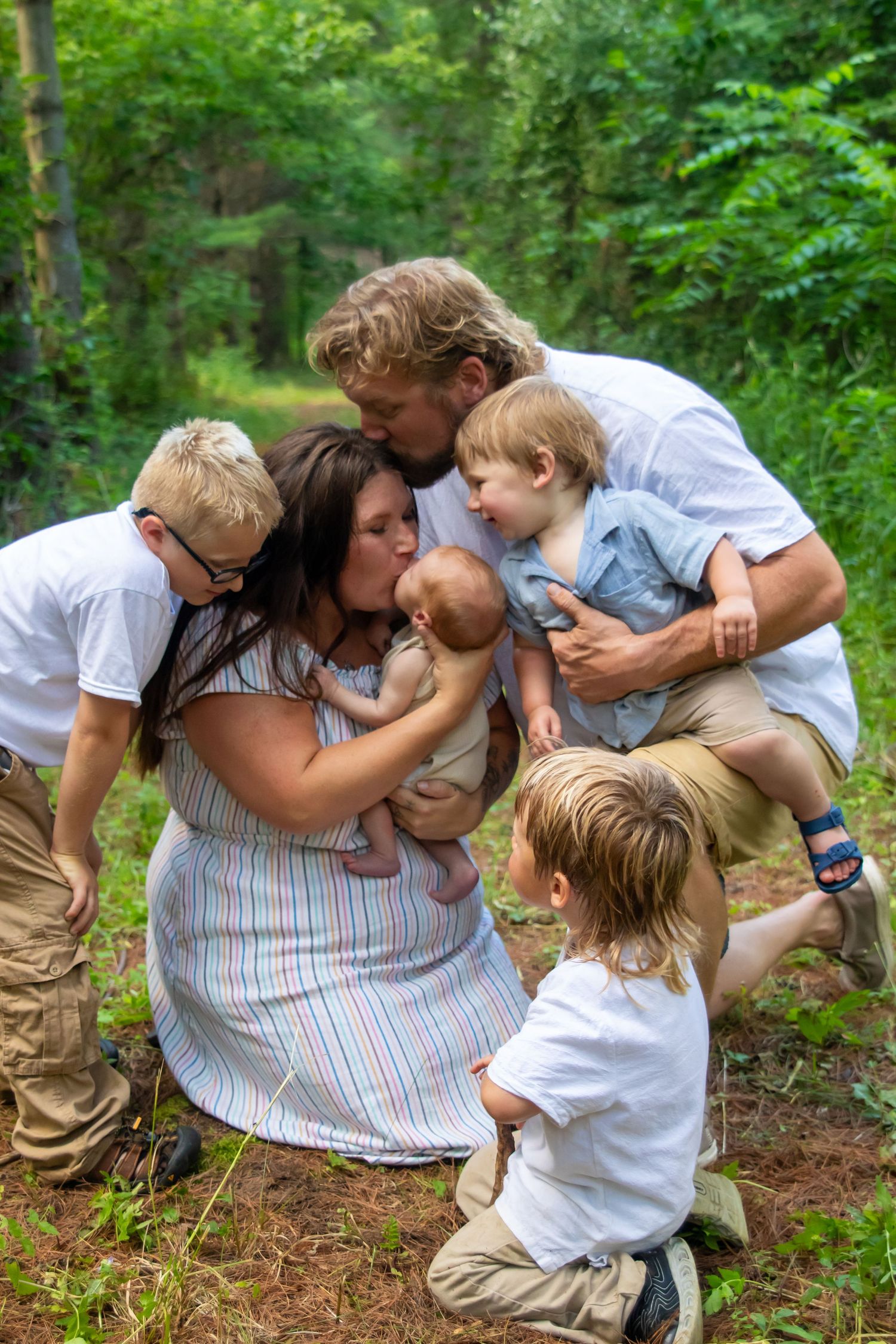 Family of five sharing kisses outdoors in a lush green forest setting, parents embracing children lovingly.
