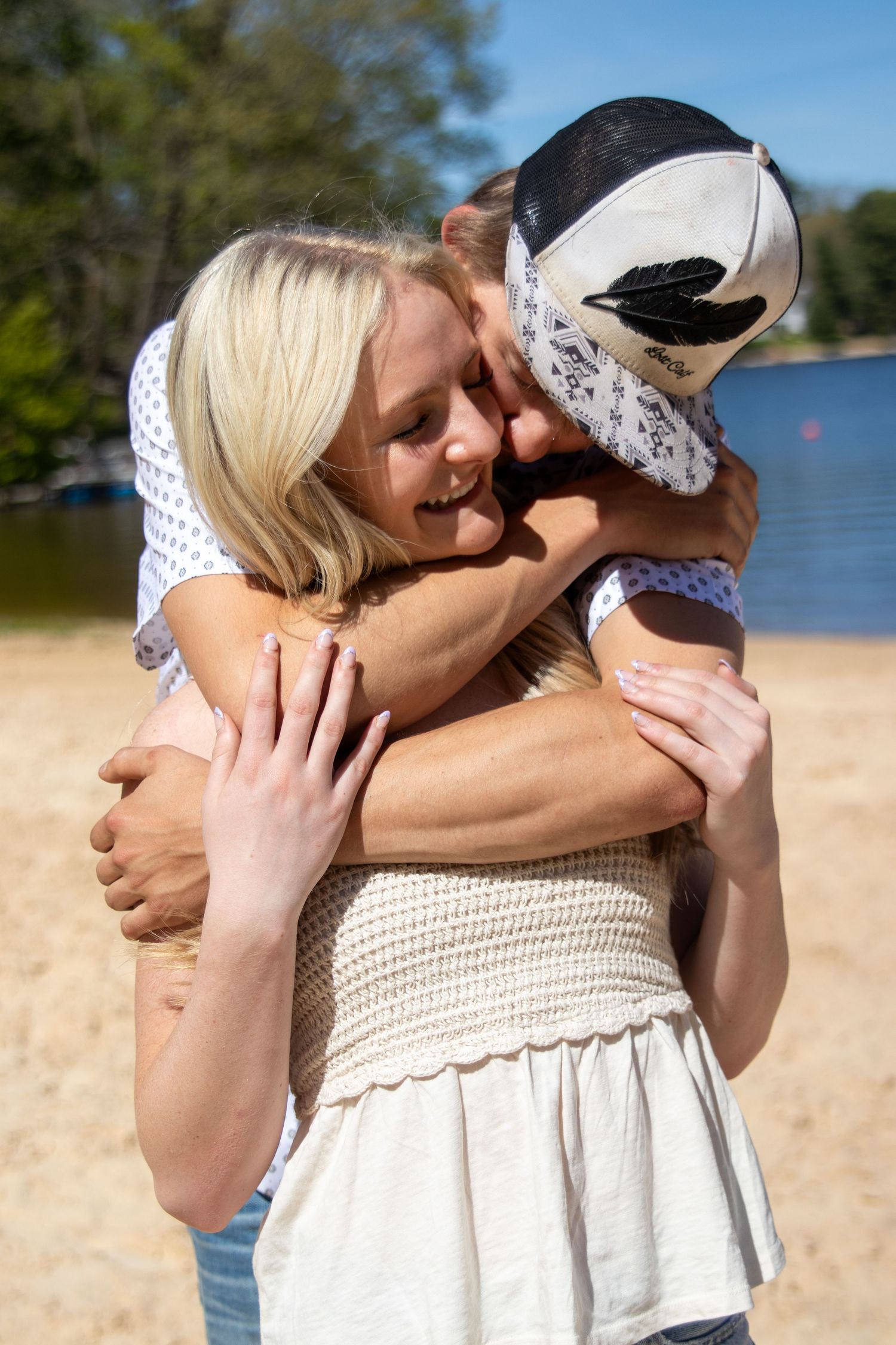 Two women sharing a warm hug on a sunny beach, one wearing a cowboy hat, with a lake and trees in the background.