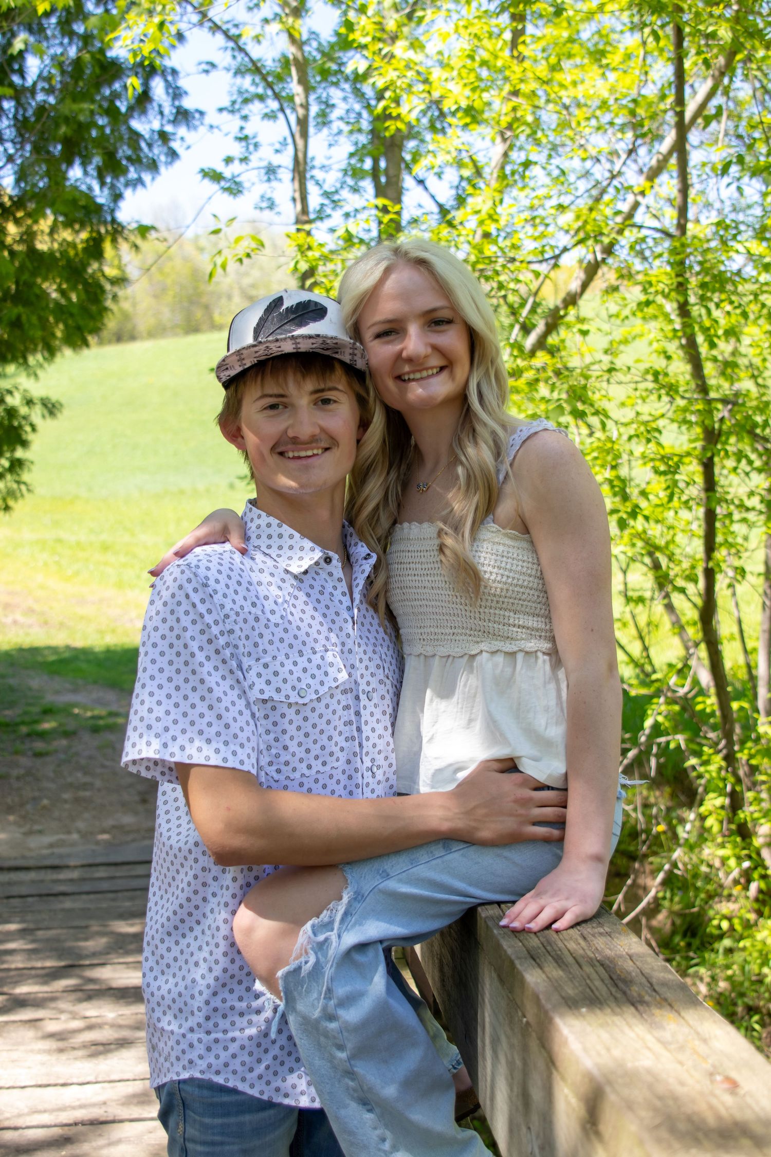 Two smiling teens pose together on a wooden bridge surrounded by lush green trees on a sunny day.