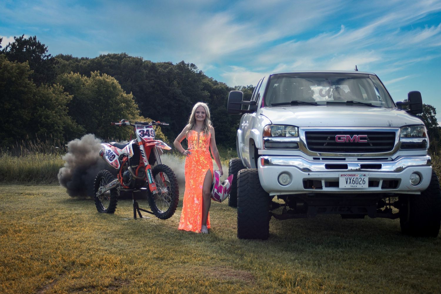 Young woman in orange prom dress posing beside GMC truck and dirt bike outdoors.