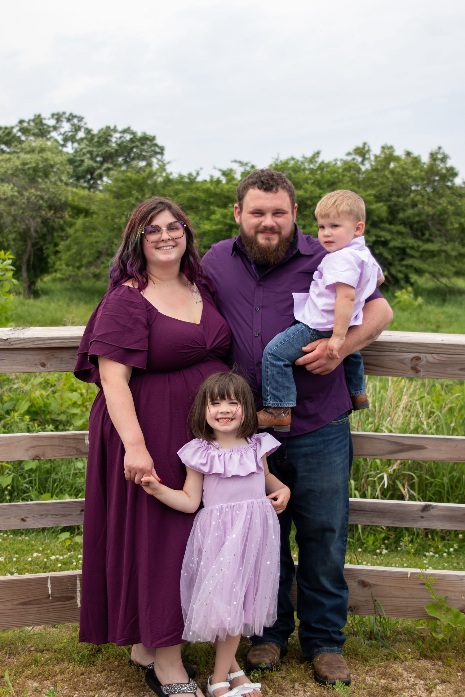 Family of four in purple outfits posing by a wooden fence with lush green trees in the background.