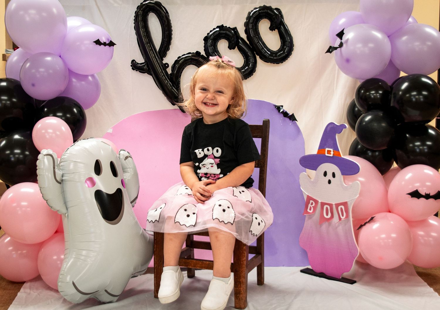 Toddler girl in BOO shirt and ghost tutu at Halloween themed photo backdrop with balloons.