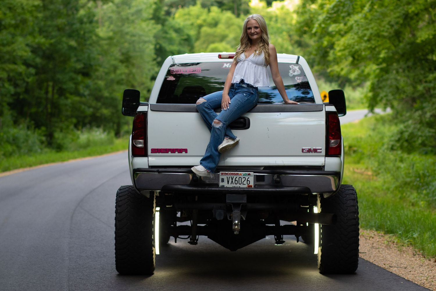 Smiling teenage girl sitting on the tailgate of a lifted white GMC truck on a tree-lined road.