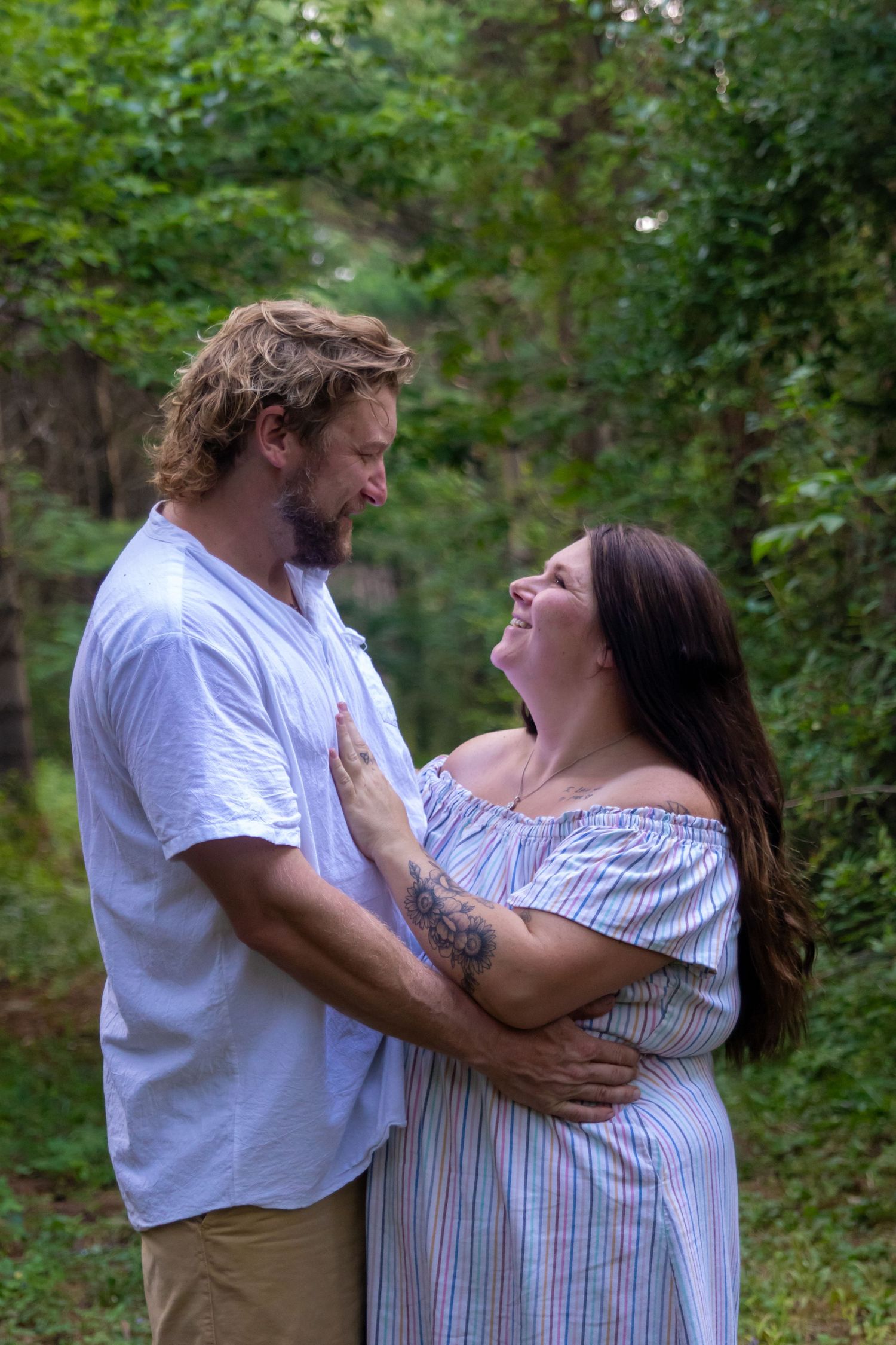 Couple sharing a tender moment outdoors, woman with tattoo in striped off-shoulder dress, man in white shirt, forest backdrop.
