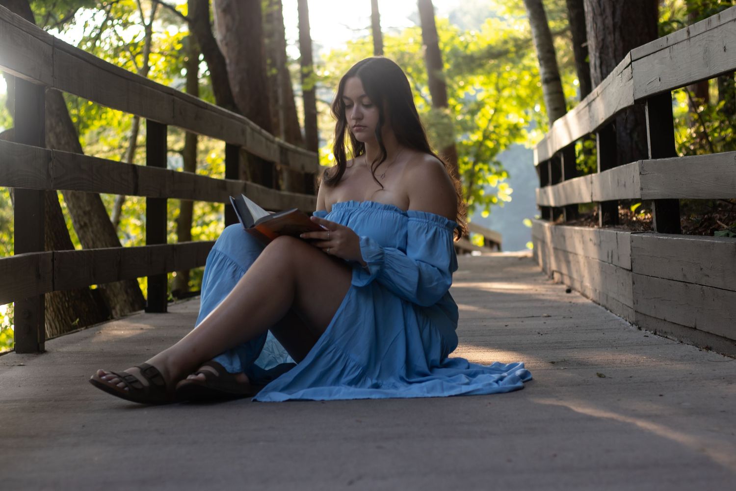 Young woman in blue off-shoulder dress reading a book while sitting on a wooden bridge path outdoors.