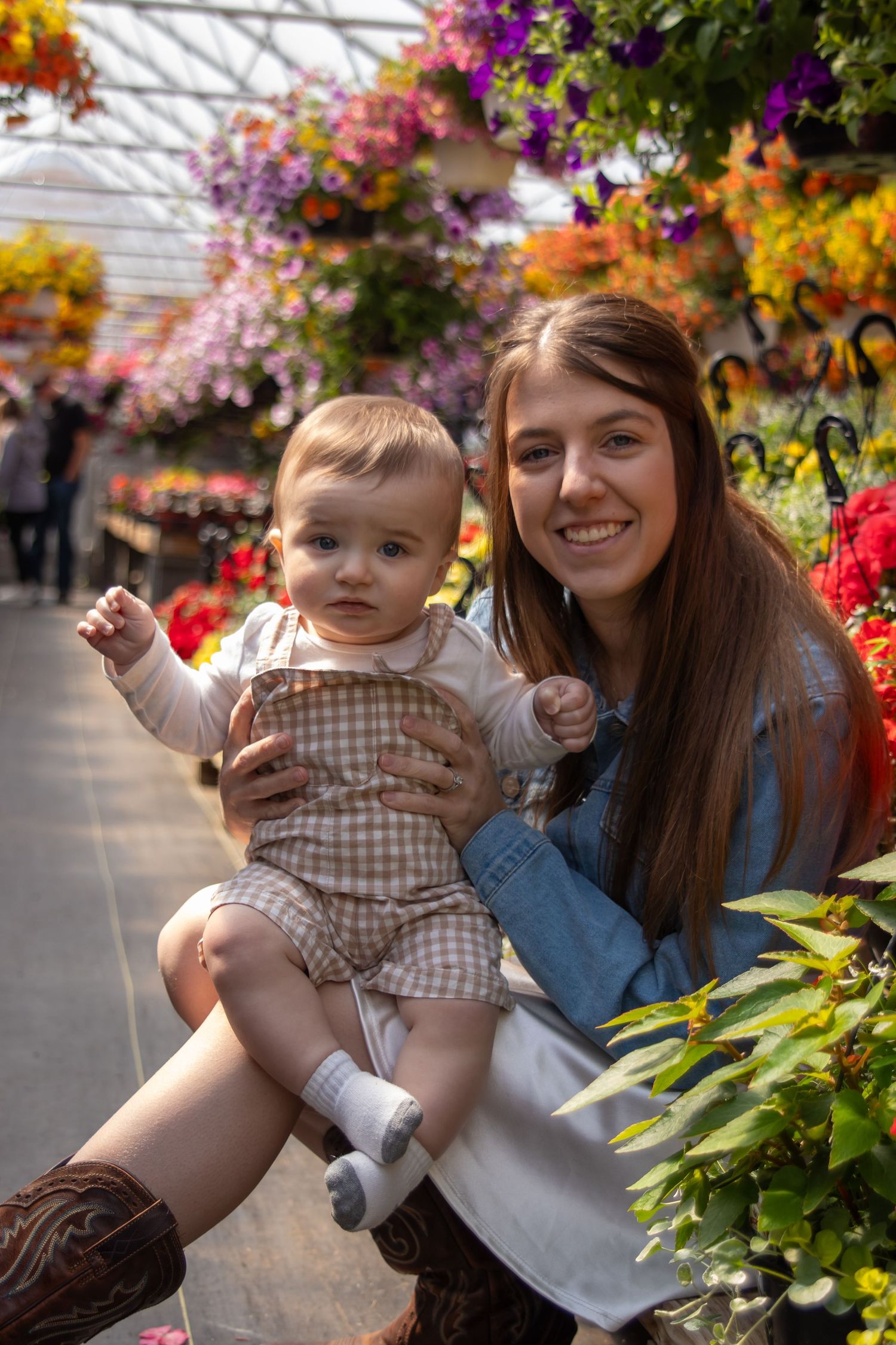 Smiling young woman holding a baby in a colorful flower greenhouse with hanging baskets.