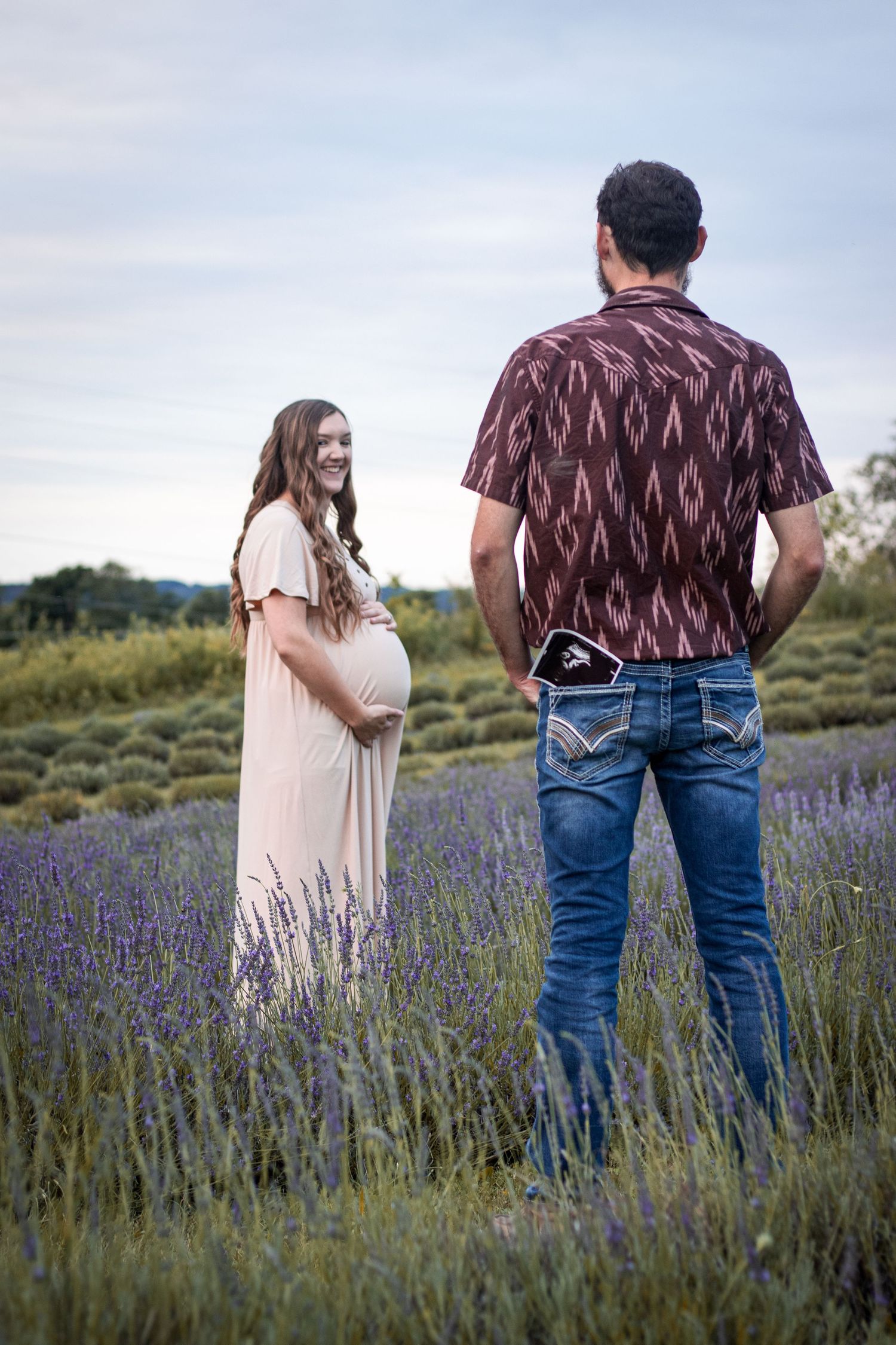 Smiling pregnant woman in blush dress stands in lavender field as partner holds ultrasound in pocket.