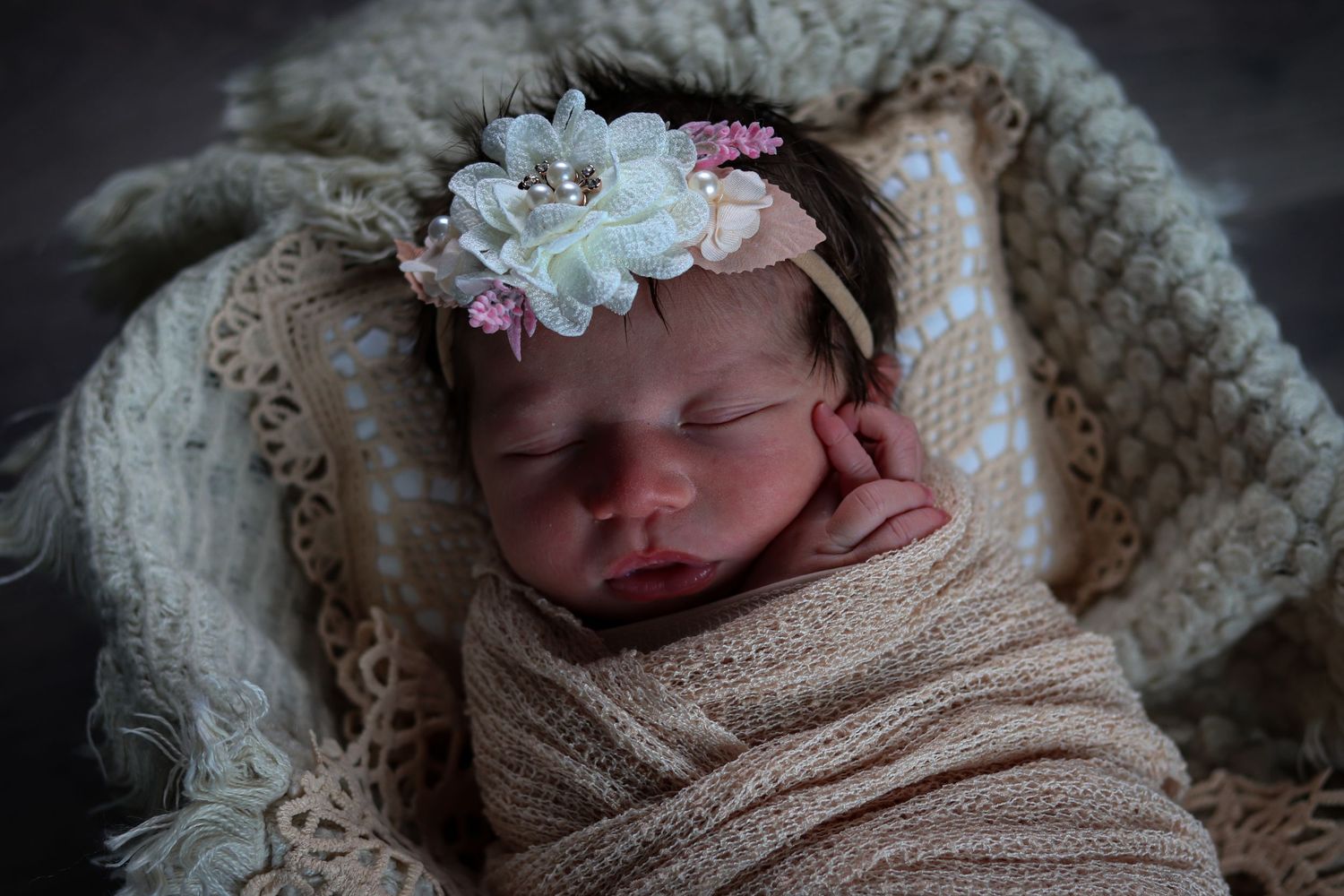 Sleeping newborn baby girl wrapped in cream swaddle wearing delicate floral headband in professional portrait.