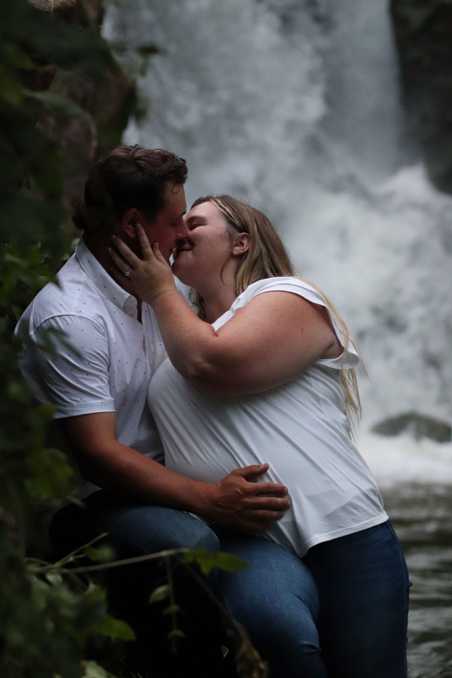 Couple shares passionate kiss in front of stunning waterfall during romantic outdoor engagement photo session.