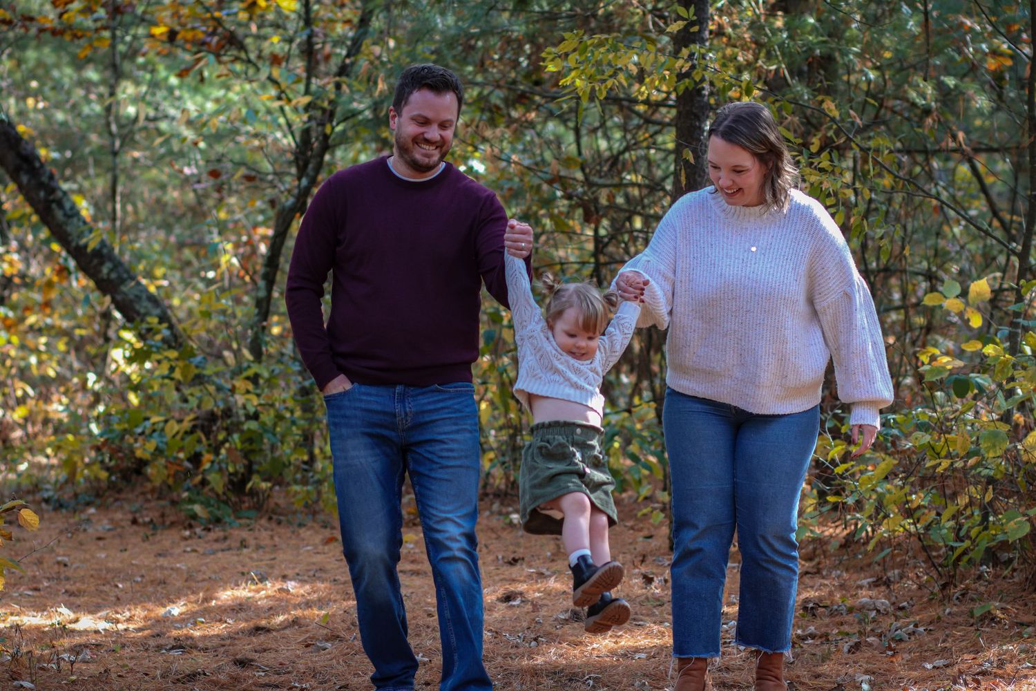 Family of three walking through autumn woods, parents swinging toddler by hands, smiling outdoors.