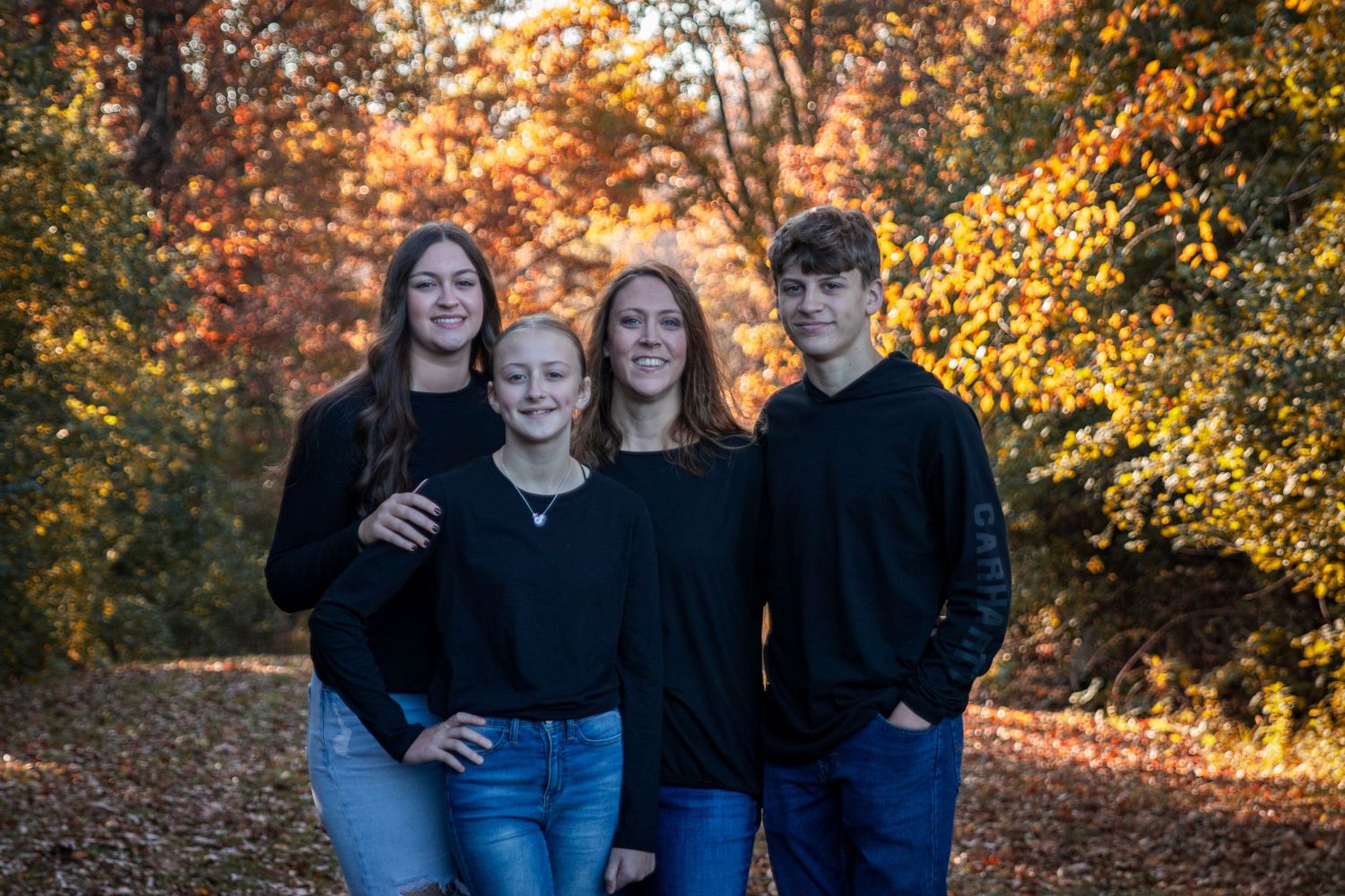 Four people in black tops pose together on a leaf-covered path surrounded by vibrant autumn foliage.