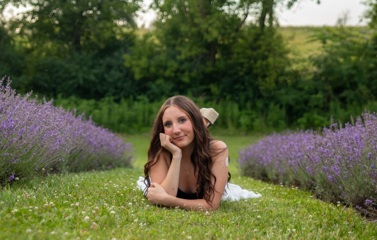 Young woman lying on grass between rows of purple lavender flowers in a lush green garden setting.