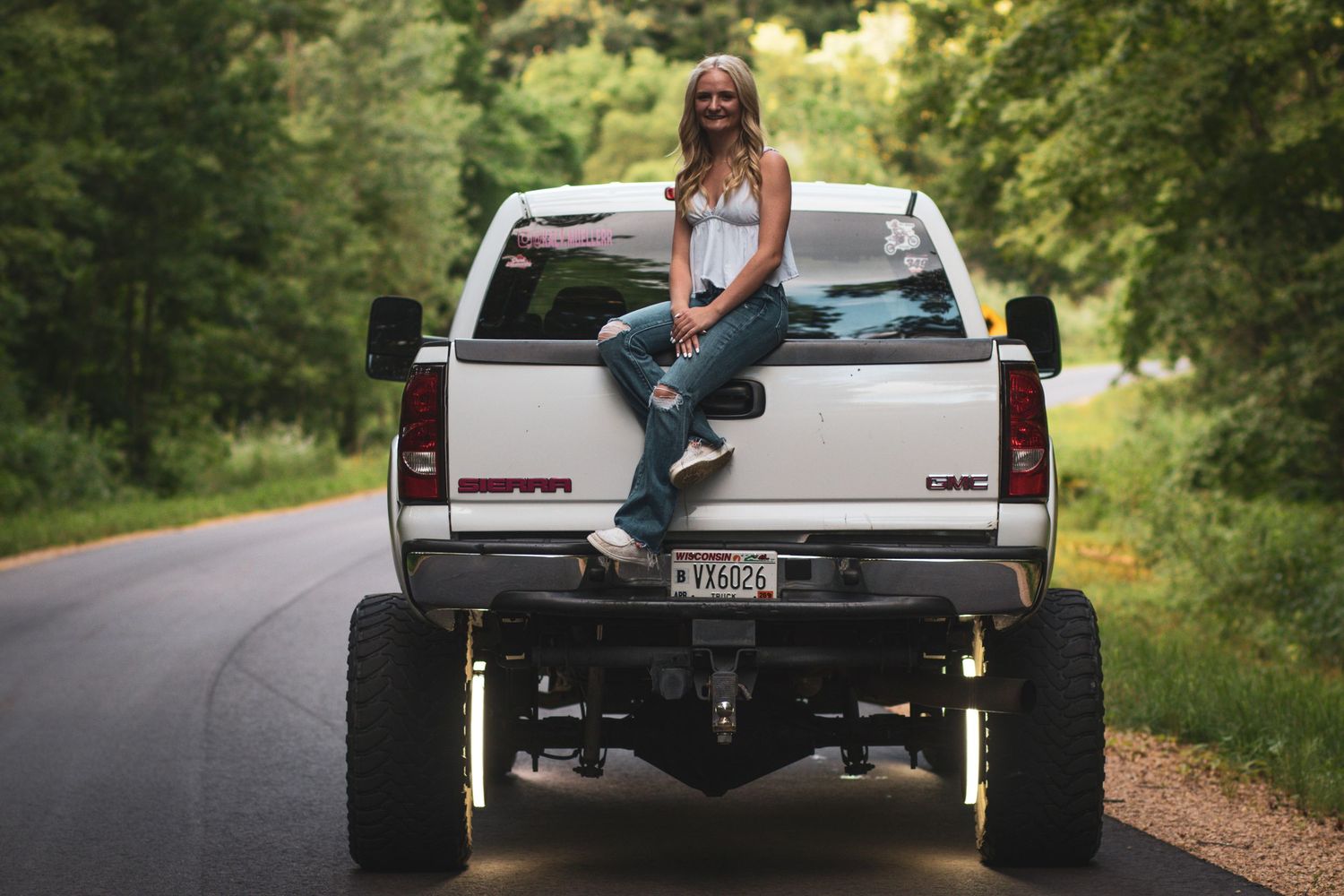 Smiling senior girl sitting on tailgate of lifted white GMC Sierra truck on tree-lined road.