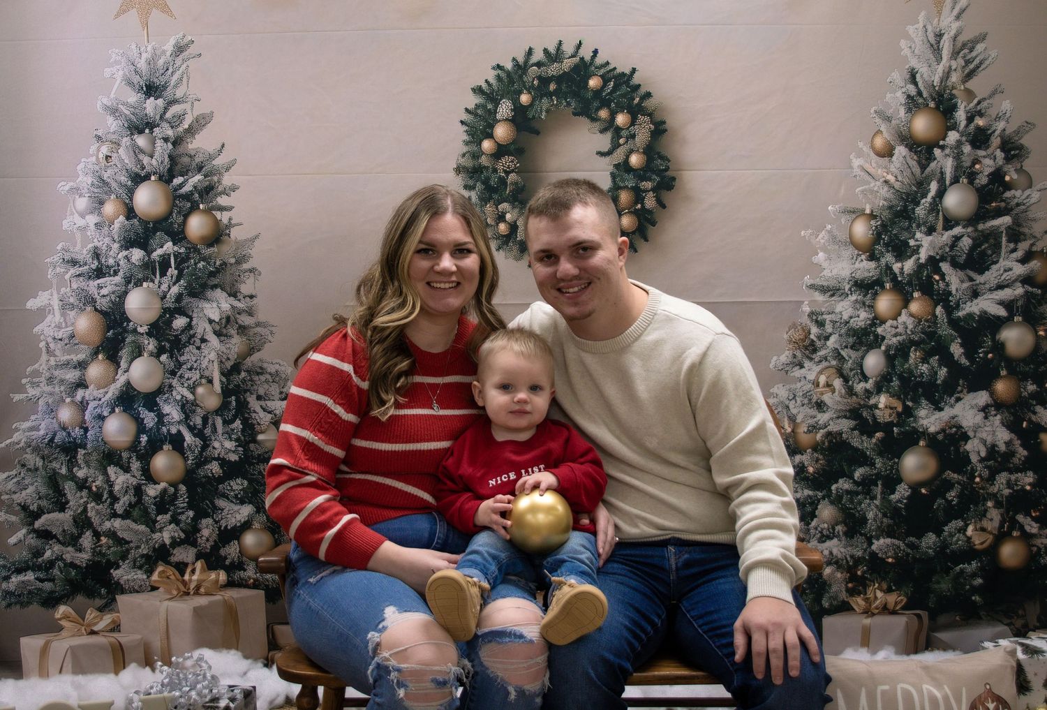 Family of three posing for Christmas portrait with decorated trees and wreath backdrop.