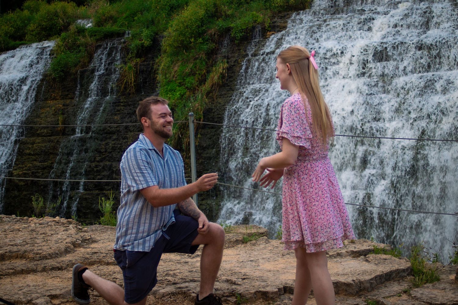 Man proposes to woman in pink dress near waterfall outdoors.