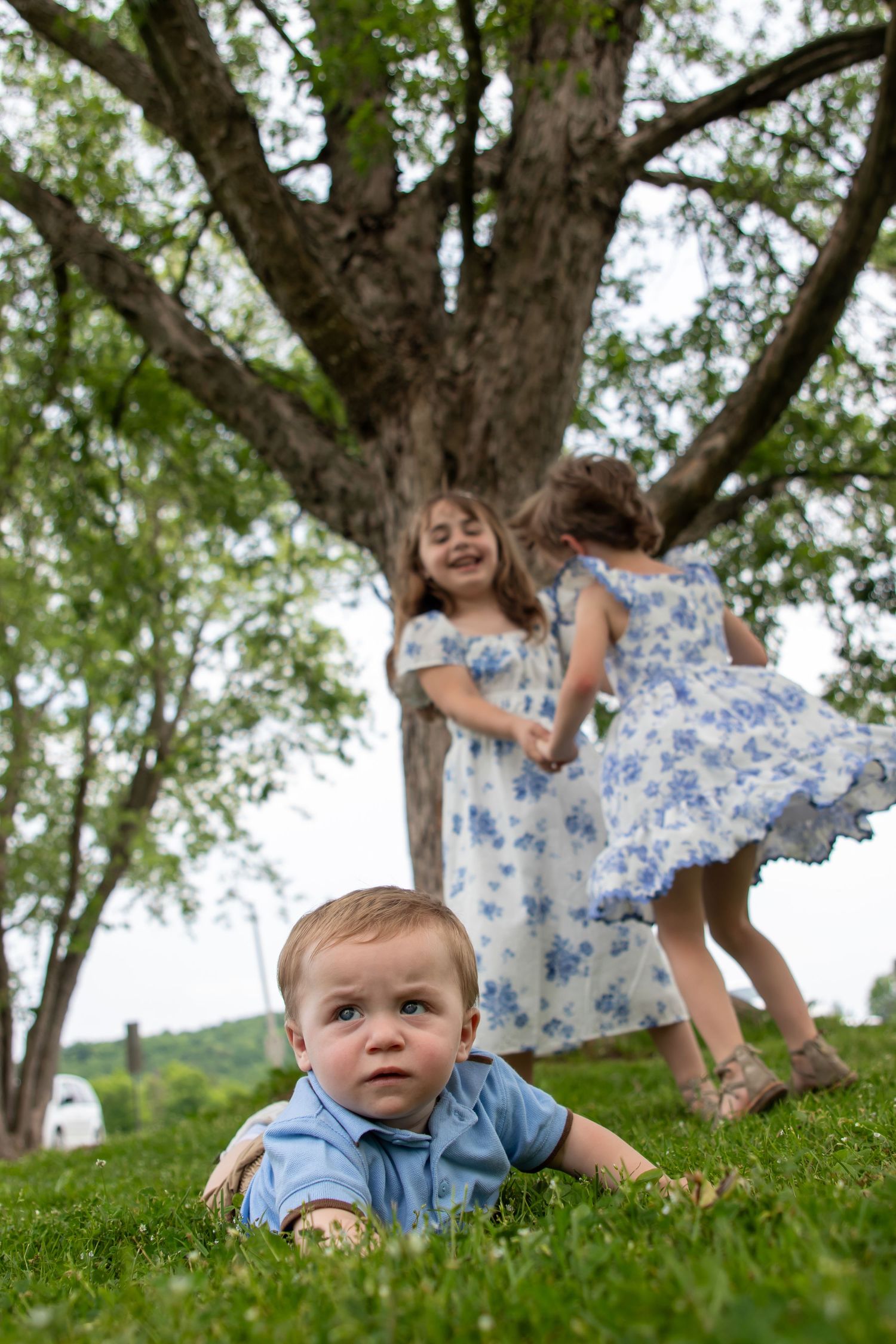 Baby boy in blue shirt crawls on grass while two girls in floral dresses play and spin under a large tree outdoors.