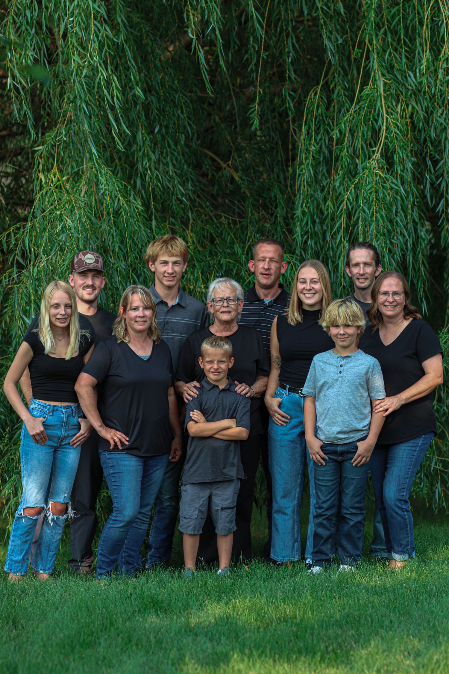 Extended family of ten dressed in black posing under a weeping willow tree outdoors.