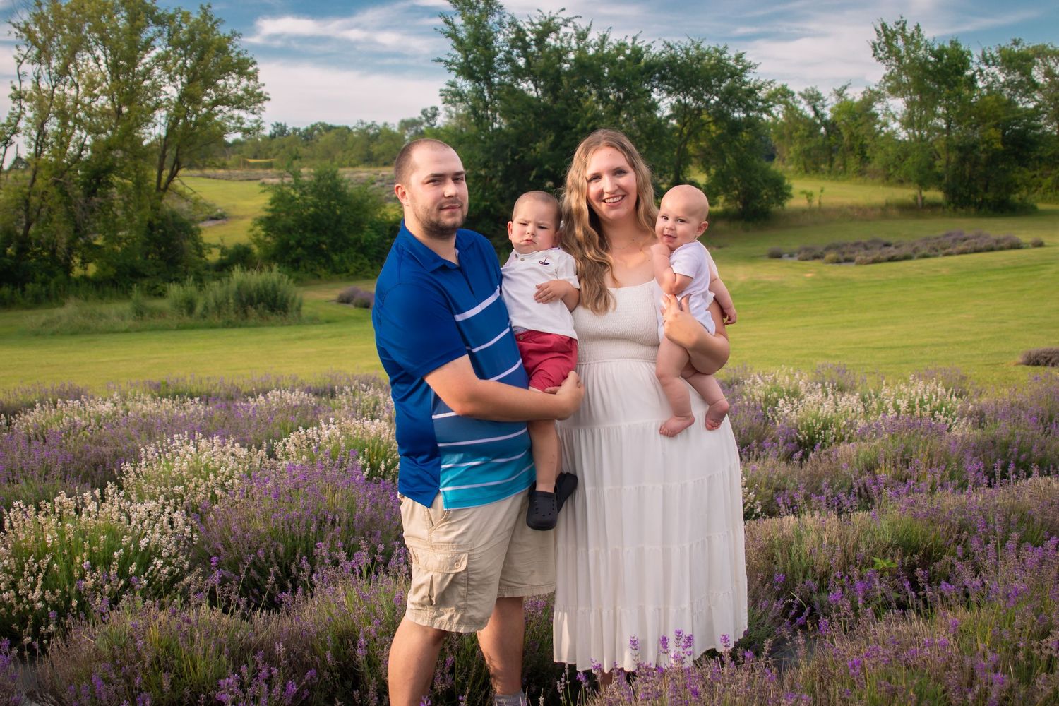 Family of four posing in a lavender field, parents holding two babies, green hills in background on a sunny day.