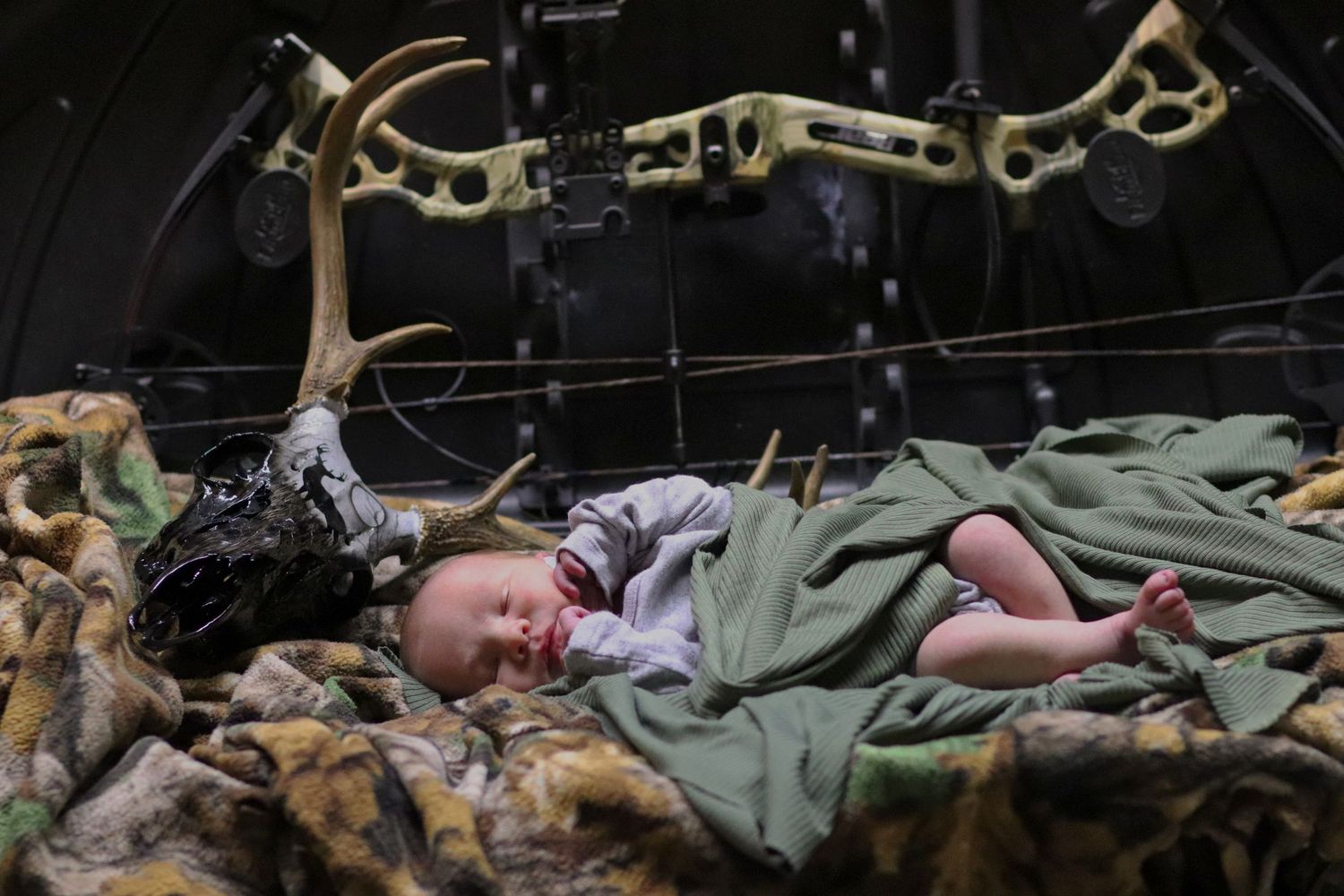 Sleeping newborn baby resting on camouflage blanket near bow and deer antlers.