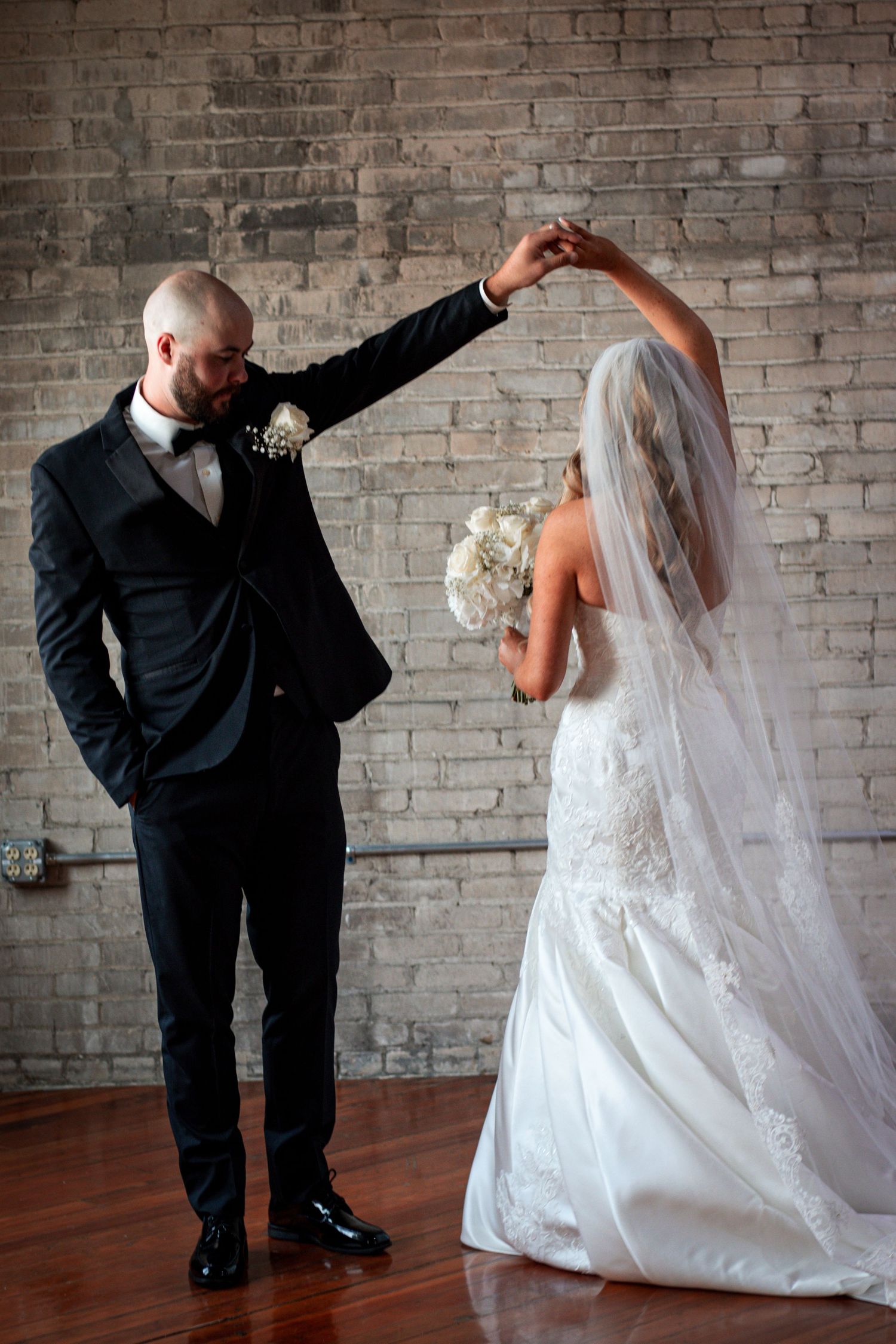 Groom in black tuxedo spinning bride in lace wedding dress in front of brick wall.