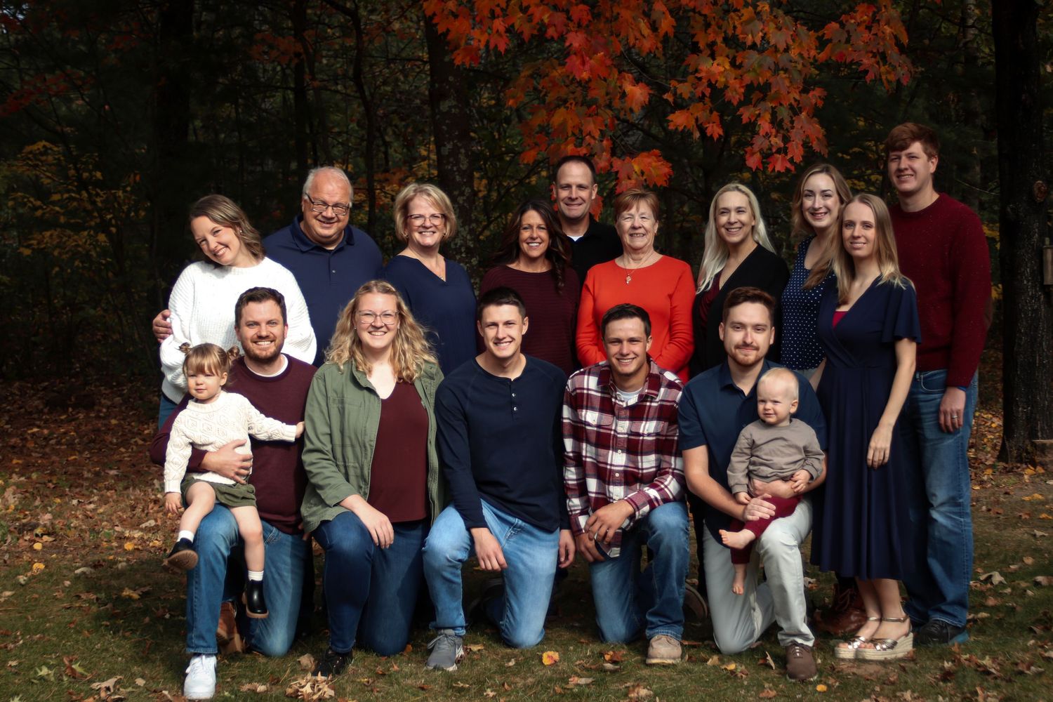 Large family group smiling together outdoors surrounded by colorful autumn foliage.