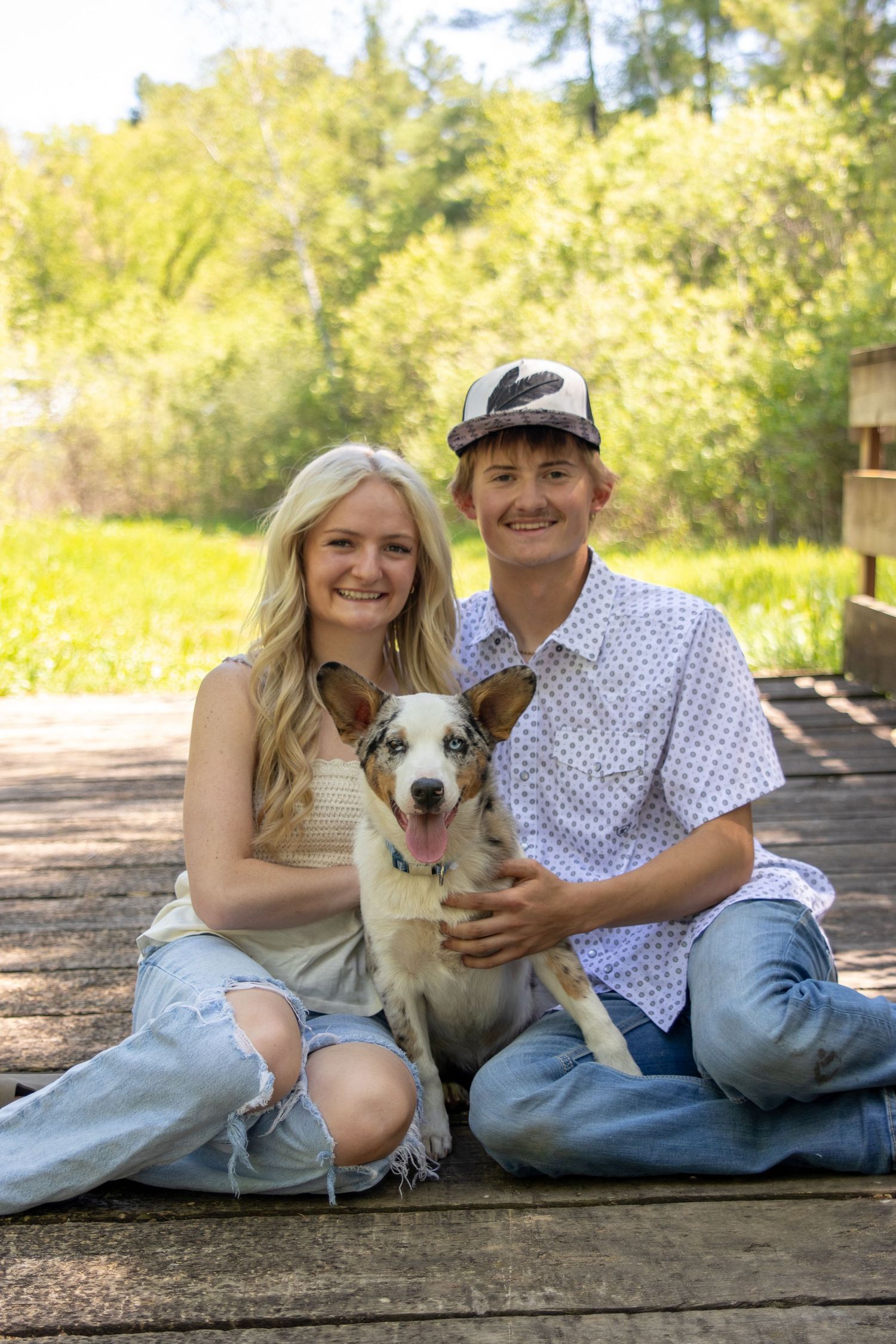 A smiling young couple sits on a wooden deck outdoors with their happy dog, surrounded by lush green trees.