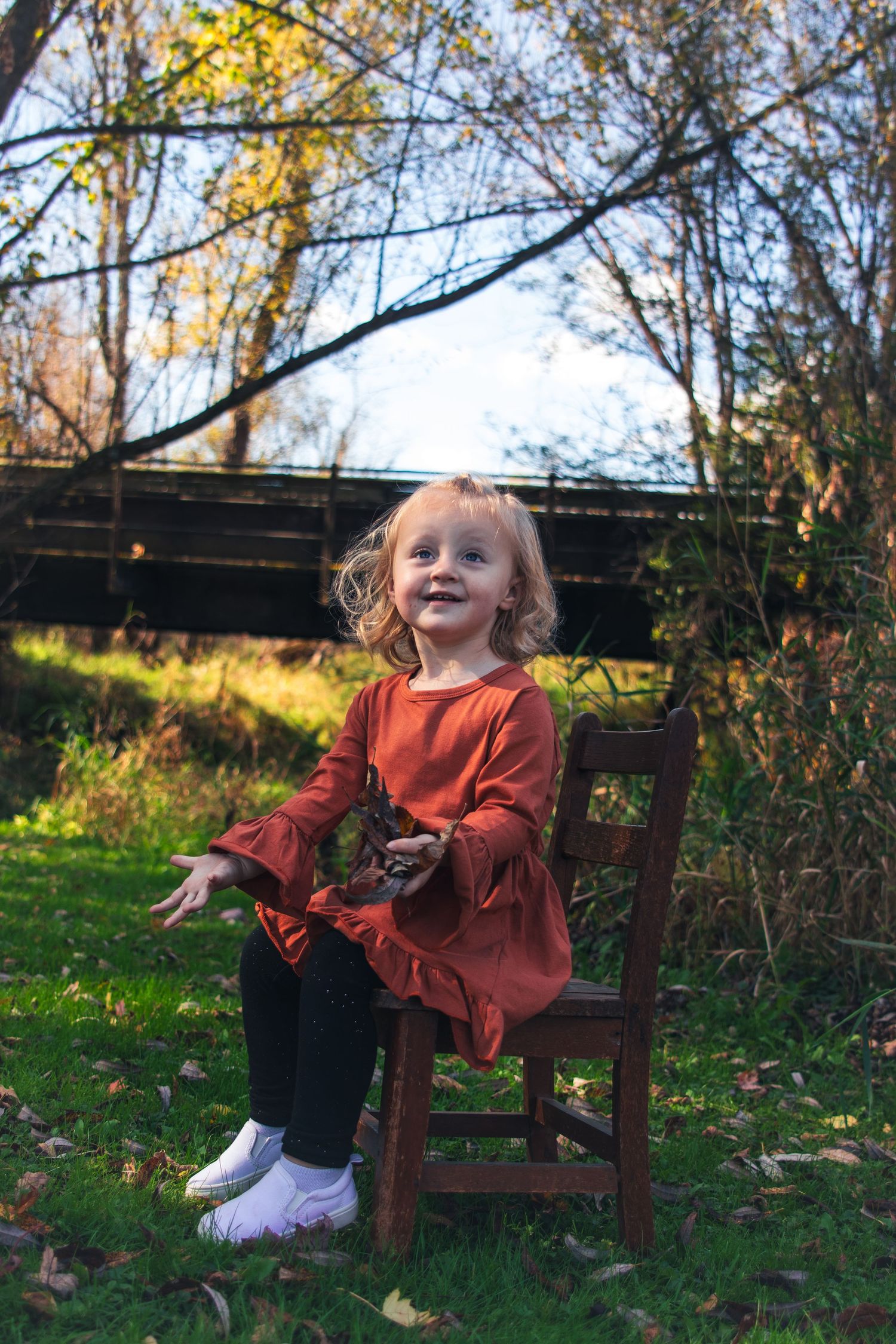 Adorable toddler girl in rust dress sitting on wooden chair outdoors in autumn.
