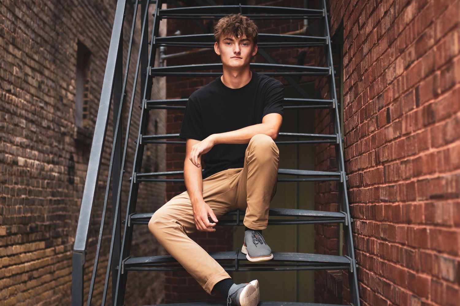 Young man in black shirt sitting on metal fire escape stairs between brick walls.