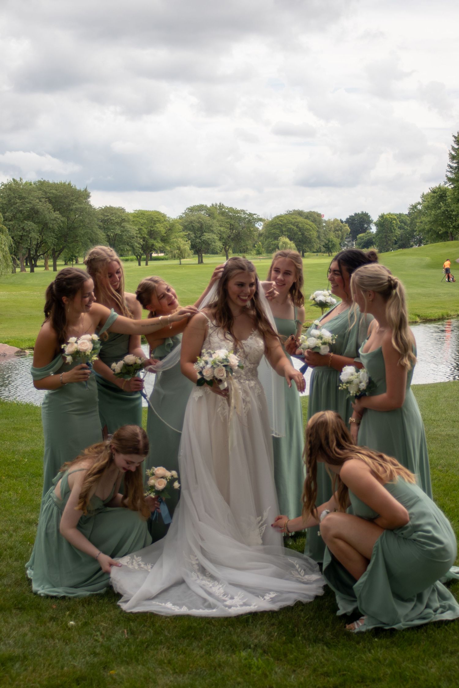 Bride surrounded by bridesmaids in sage green gowns holding white bouquets on a lush golf course.