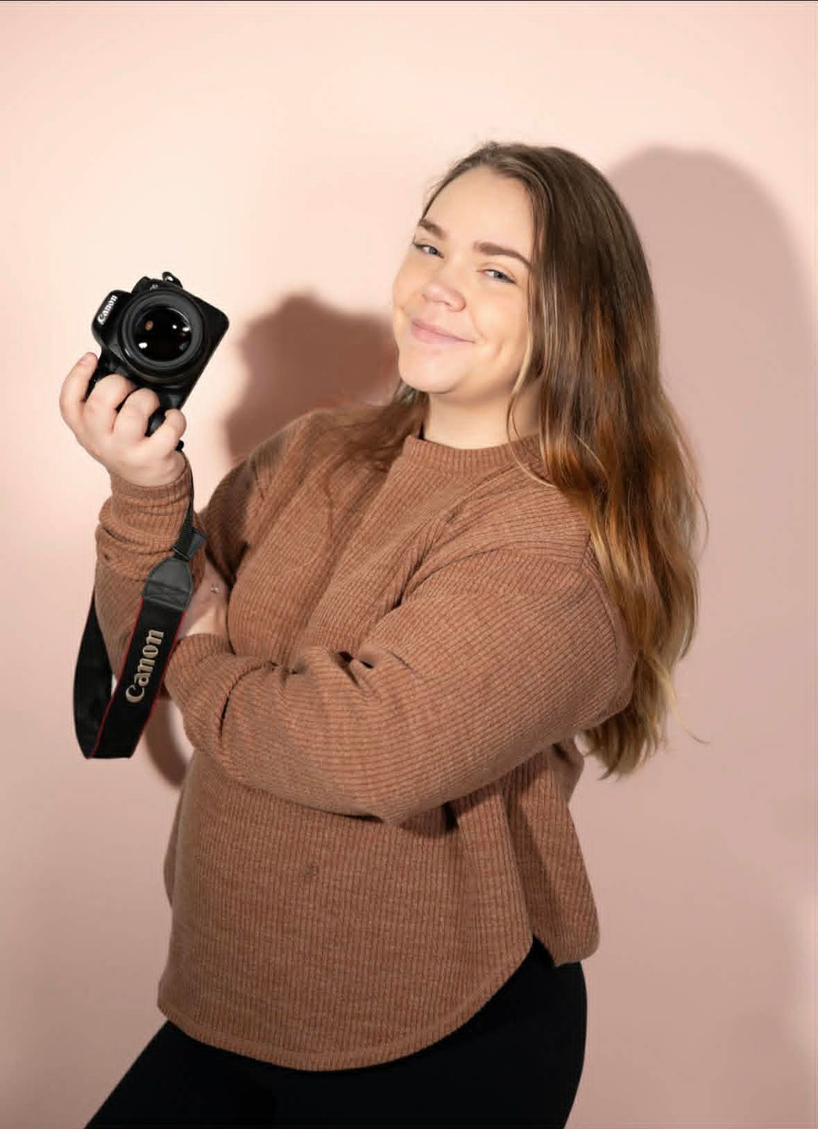Young woman in brown knit sweater holding Canon DSLR camera and smiling on pink background.