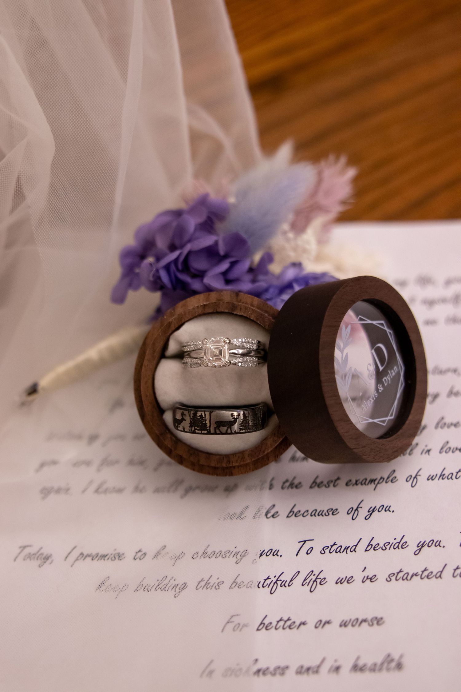 Wedding rings in a wooden ring box on handwritten vows, with a floral bouquet and bridal veil backdrop.