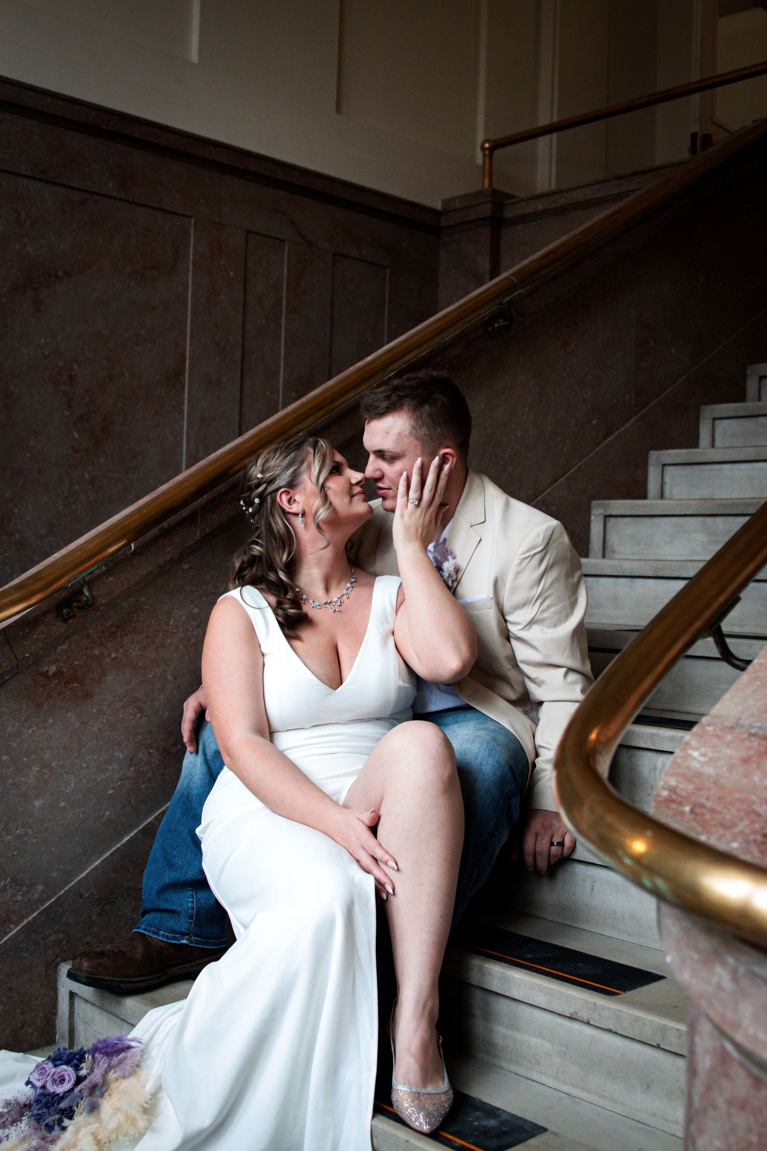 Bride and groom share a romantic moment sitting on marble stairs, bride in white gown, groom in tan suit.