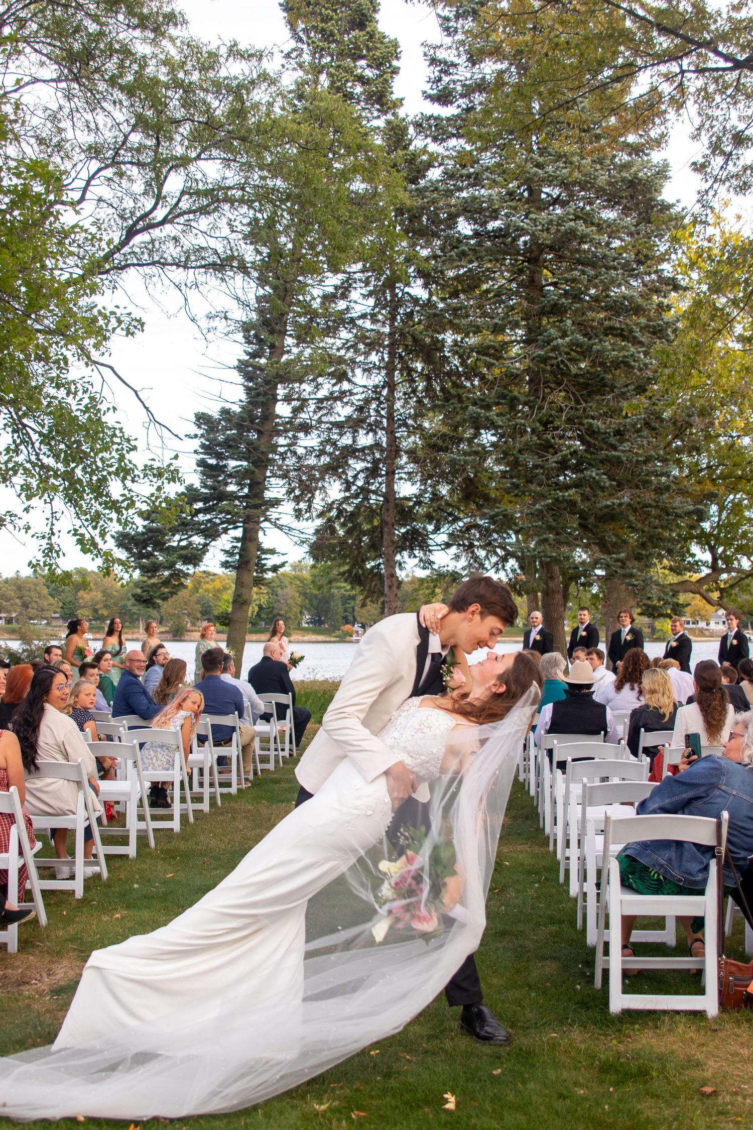 Groom dips bride in a kiss at outdoor wedding ceremony with white chairs and tall pine trees.