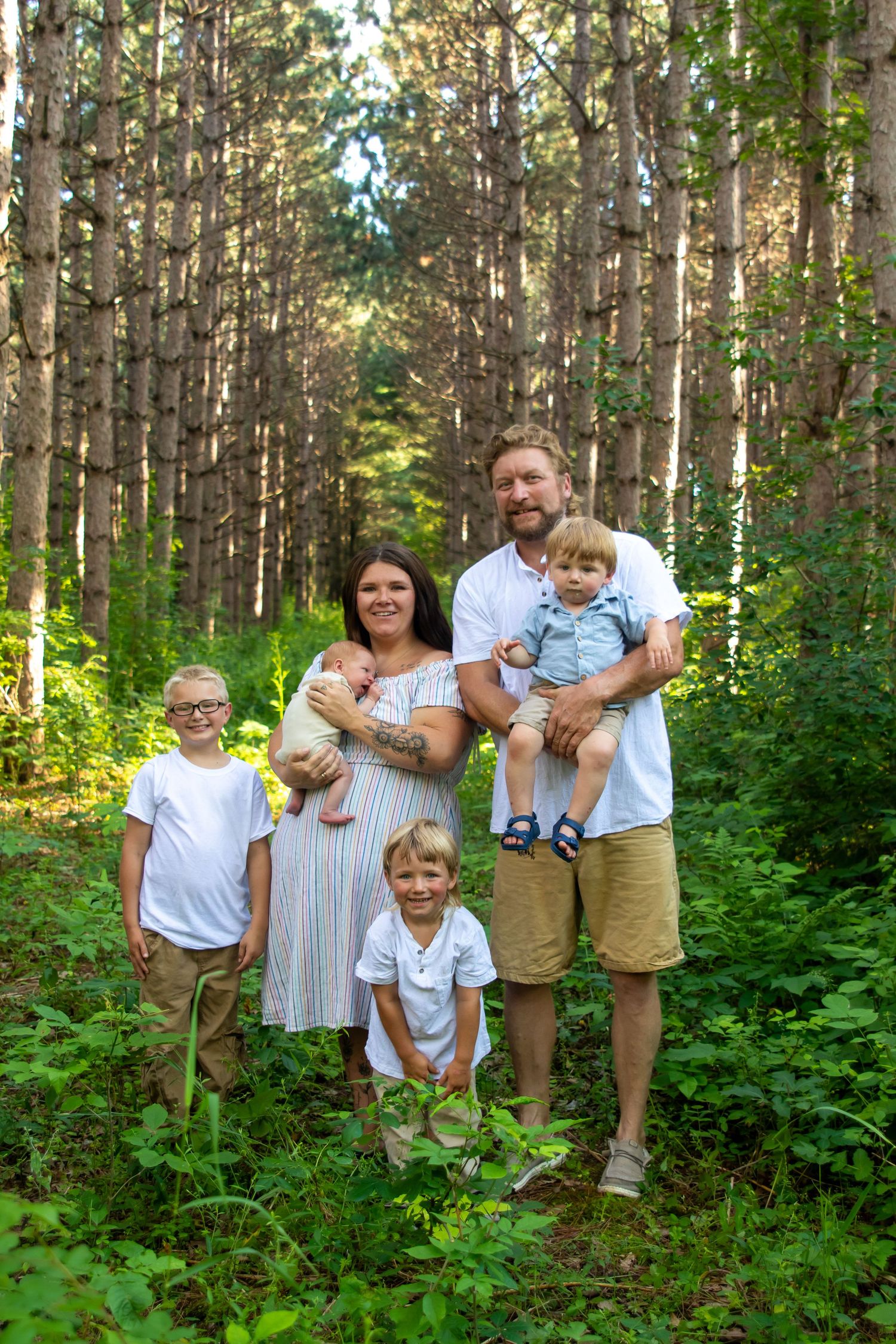 Family of six posing in a lush green forest, parents holding young children, wearing casual white and tan outfits.