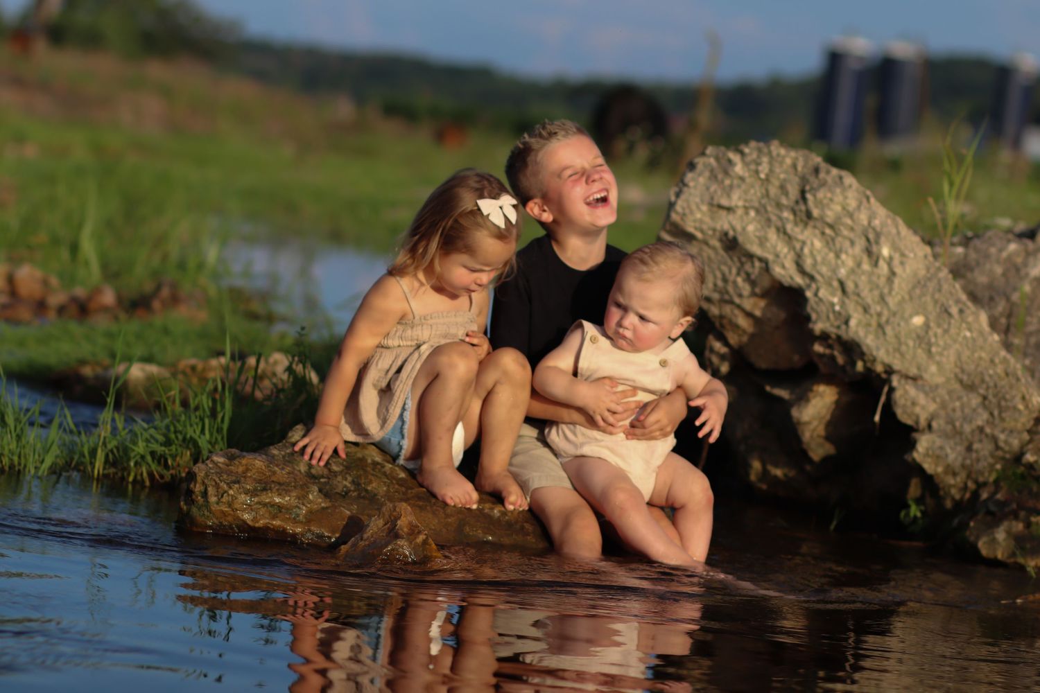 Three happy children laughing and sitting on rocks beside a calm rural pond in golden light.