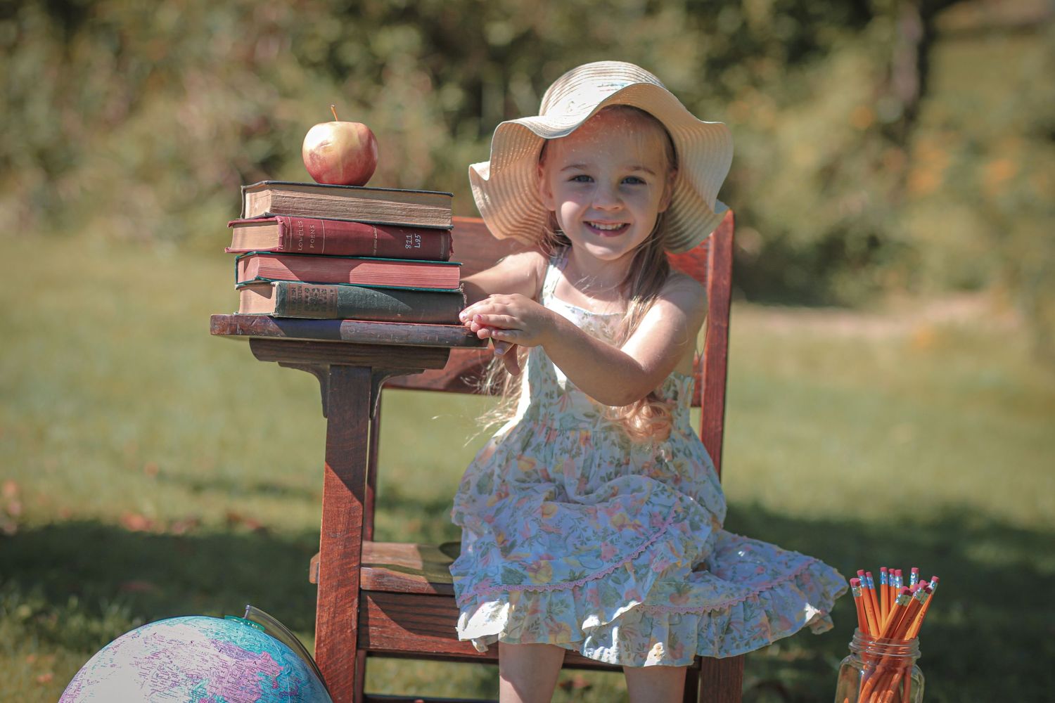 Adorable little girl in floral dress and sun hat smiles beside stack of books for back to school portrait.
