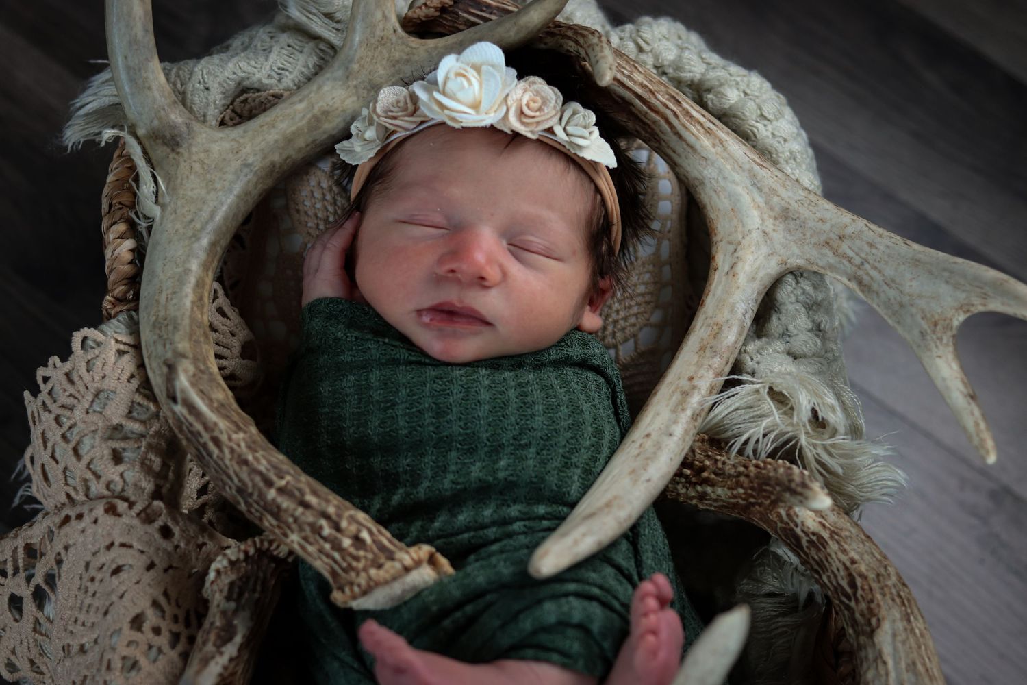 Newborn baby sleeping in antler basket wrapped in green swaddle with floral headband.