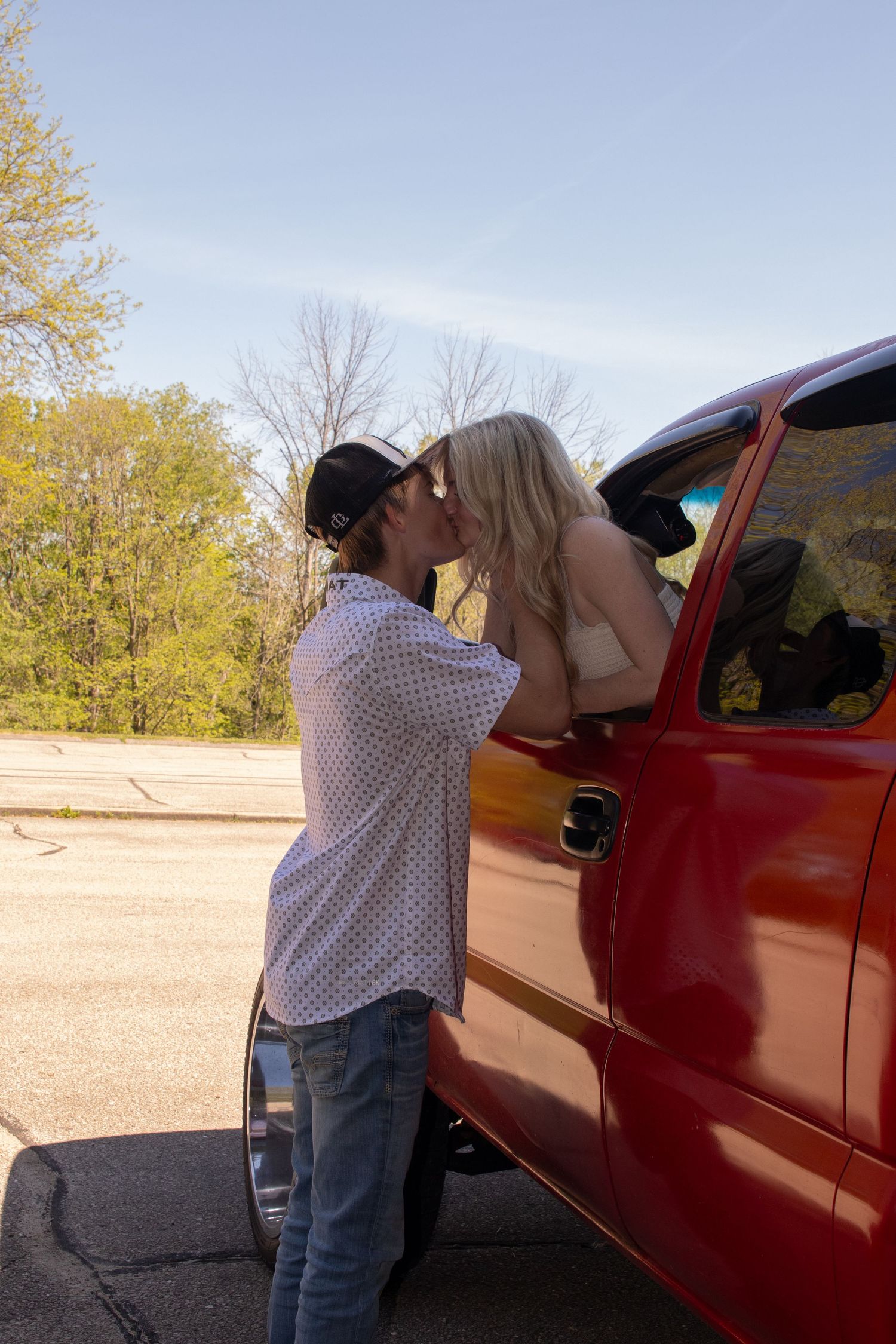 Couple sharing a kiss beside a red pickup truck in a sunny parking lot with trees in the background.