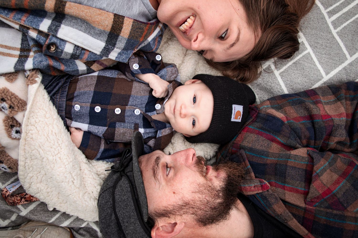 Young family of three lying together smiling with baby in Carhartt beanie on cozy blanket.