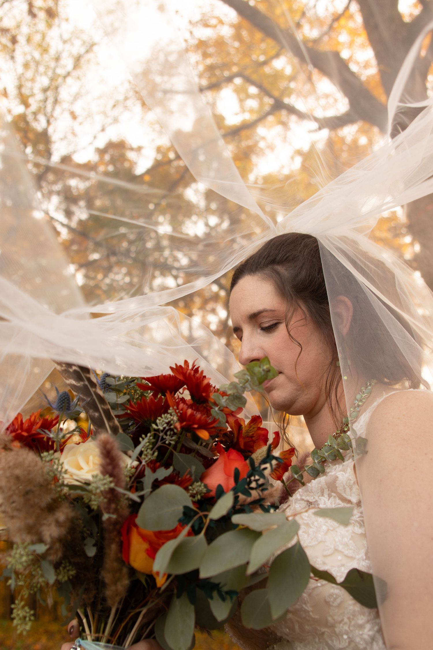 Bride veiled in tulle gazes down at a vibrant autumn bouquet of red and orange flowers.