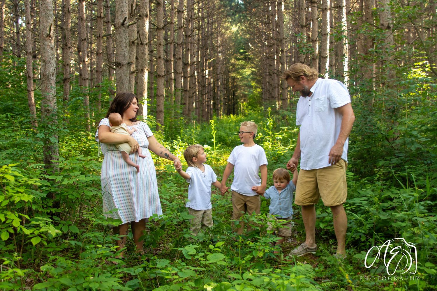 Family of six poses in a lush green forest, parents holding children among tall trees in a bright outdoor setting.