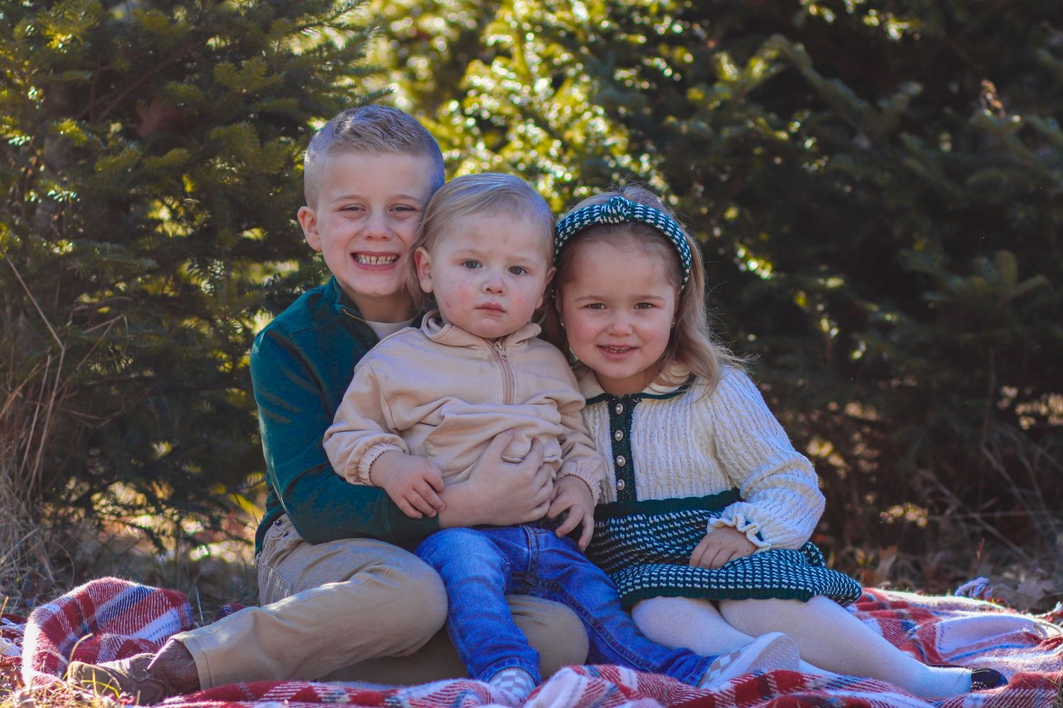 Three young siblings pose together on red plaid blanket in front of evergreen trees for family portrait.