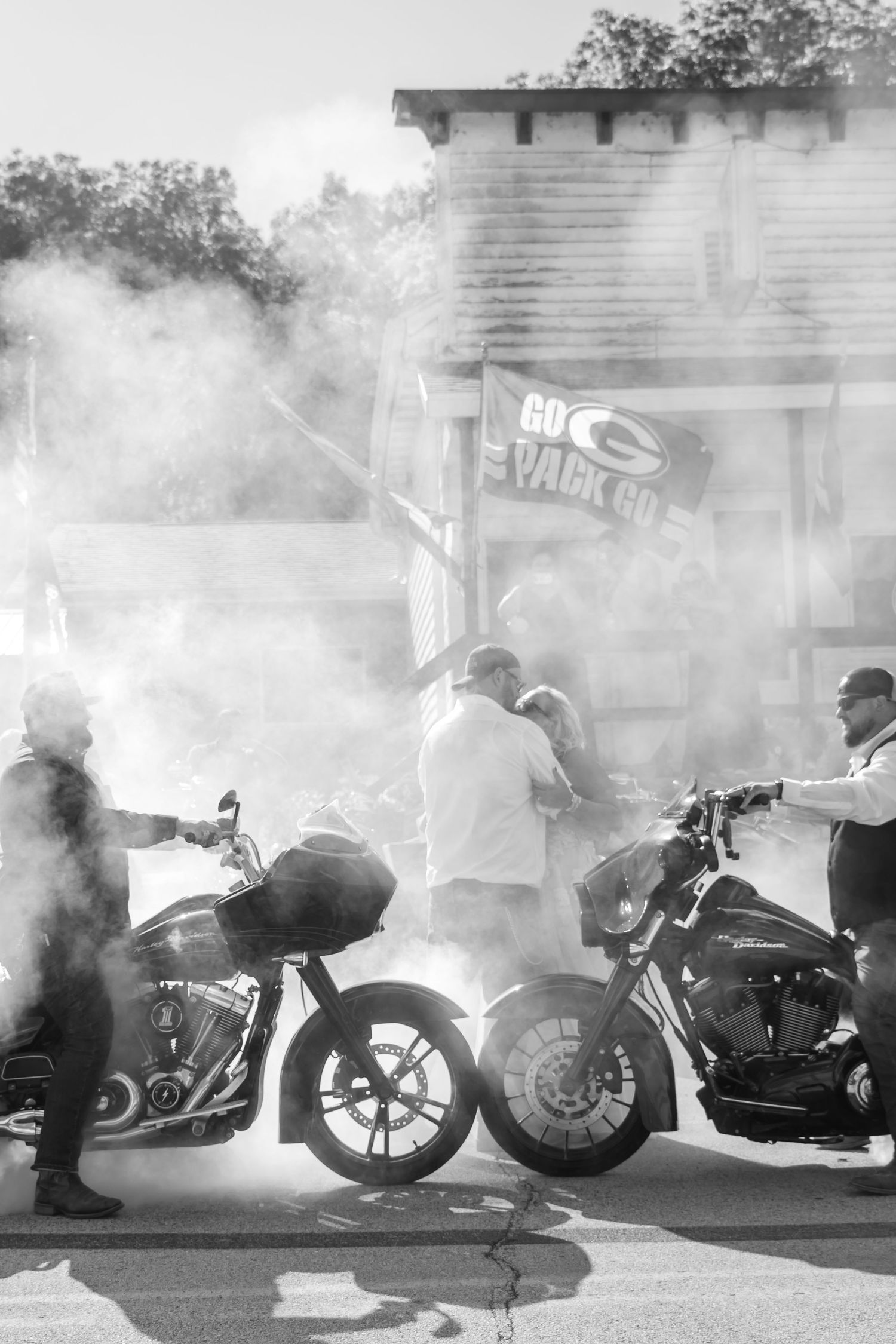 Black and white photo of bikers with Harley-Davidson motorcycles surrounded by smoke near a Green Bay Packers flag.